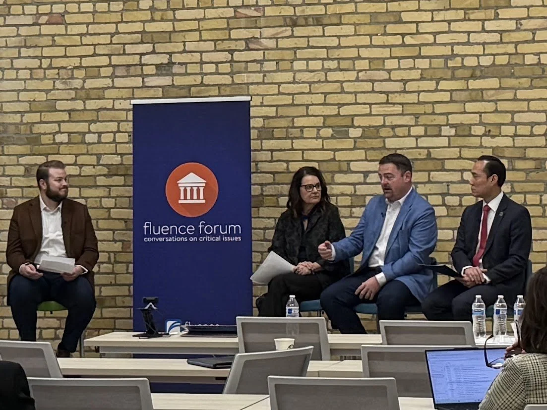 Four people seated on stage in front of a brick wall during a panel discussion. There is a banner with a logo and text that says 'fluency forum conversations on critical issues.' Water bottles are placed on the table in front of the panelists, and a laptop and water bottles are seen on the tables in the audience.