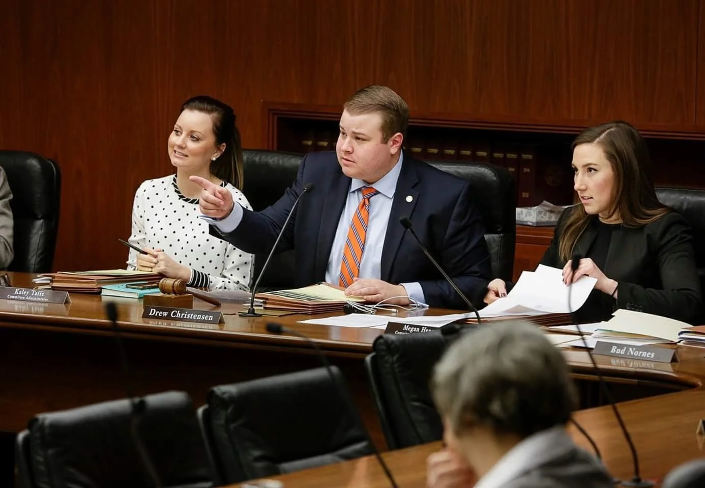 Three people sit at a conference table during a meeting, with a man in a suit pointing and speaking, and two women listening, one holding papers.