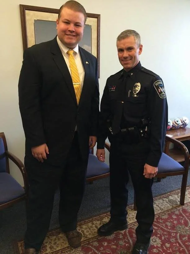 Two men, one in a suit with a yellow striped tie and the other in a police uniform, are standing side by side and smiling for the photo inside a room with chairs and a patterned rug.