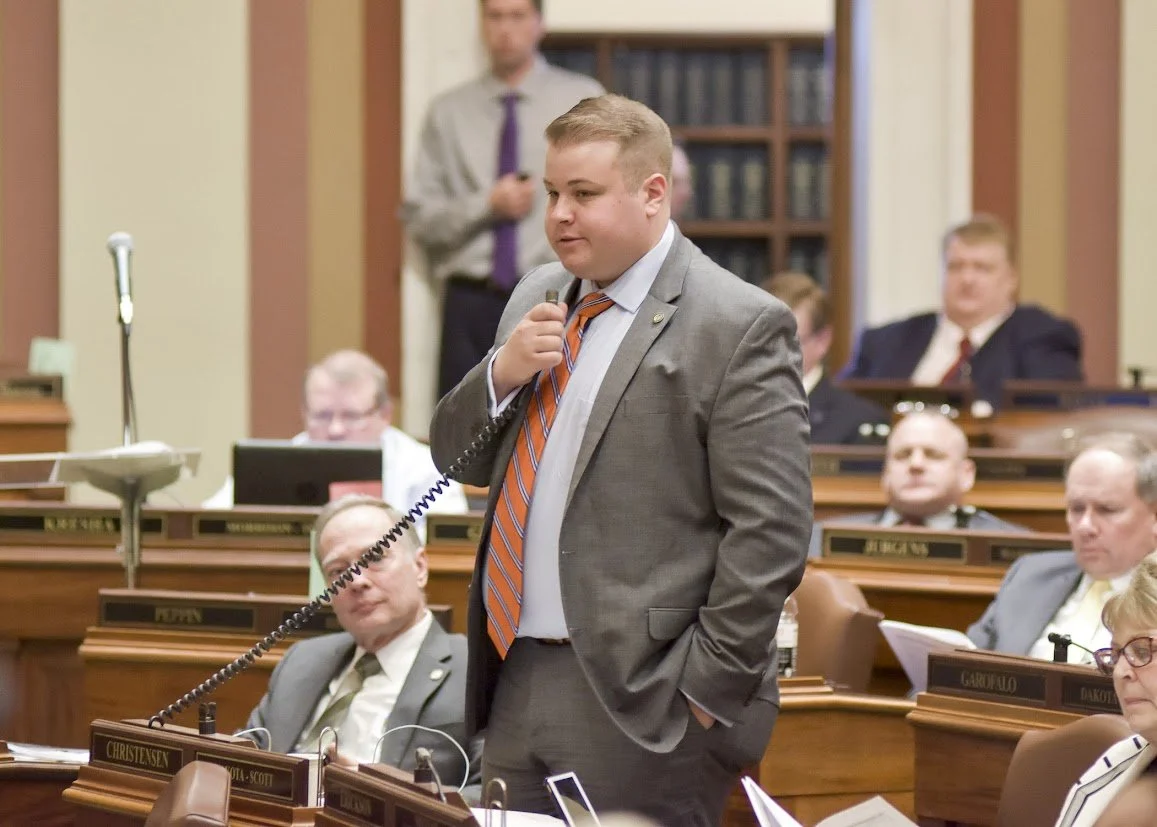 A young man in a gray suit, orange striped tie, and white shirt standing in a legislative chamber, holding a microphone near his face with his right hand, while his left hand is in his pocket. Several others are seated or standing behind him, with nameplates on the desks.