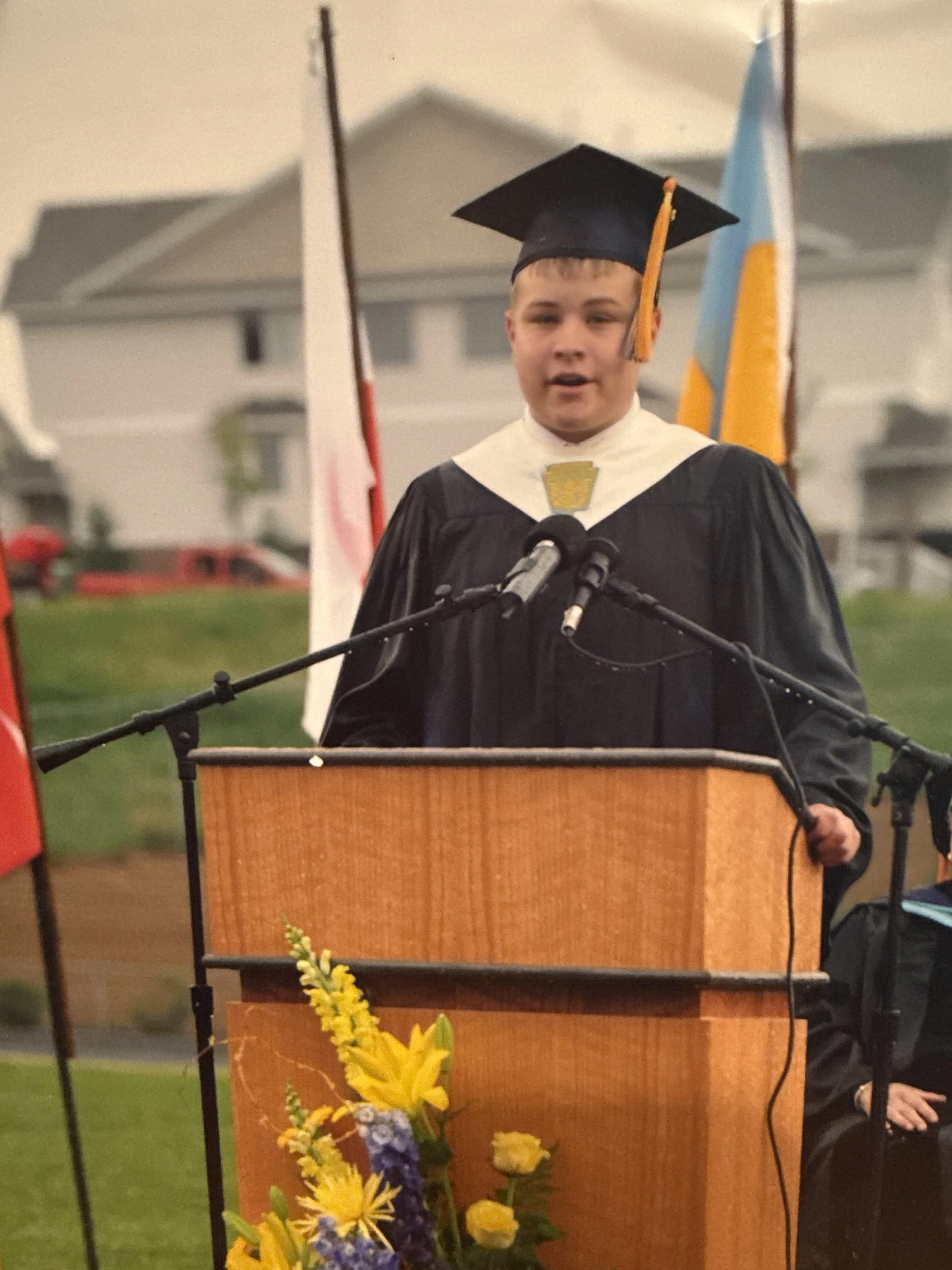 A young boy in a graduation cap and gown giving a speech at an outdoor ceremony, standing behind a podium decorated with yellow flowers.
