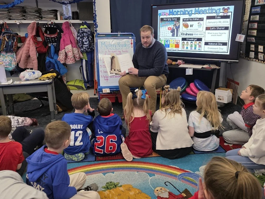 A teacher reads a book to a group of young students sitting on a colorful rainbow-themed rug in a classroom. The classroom has jackets and backpacks hanging on hooks, and a whiteboard with a presentation titled 'Morning Meeting' displayed on a large screen.
