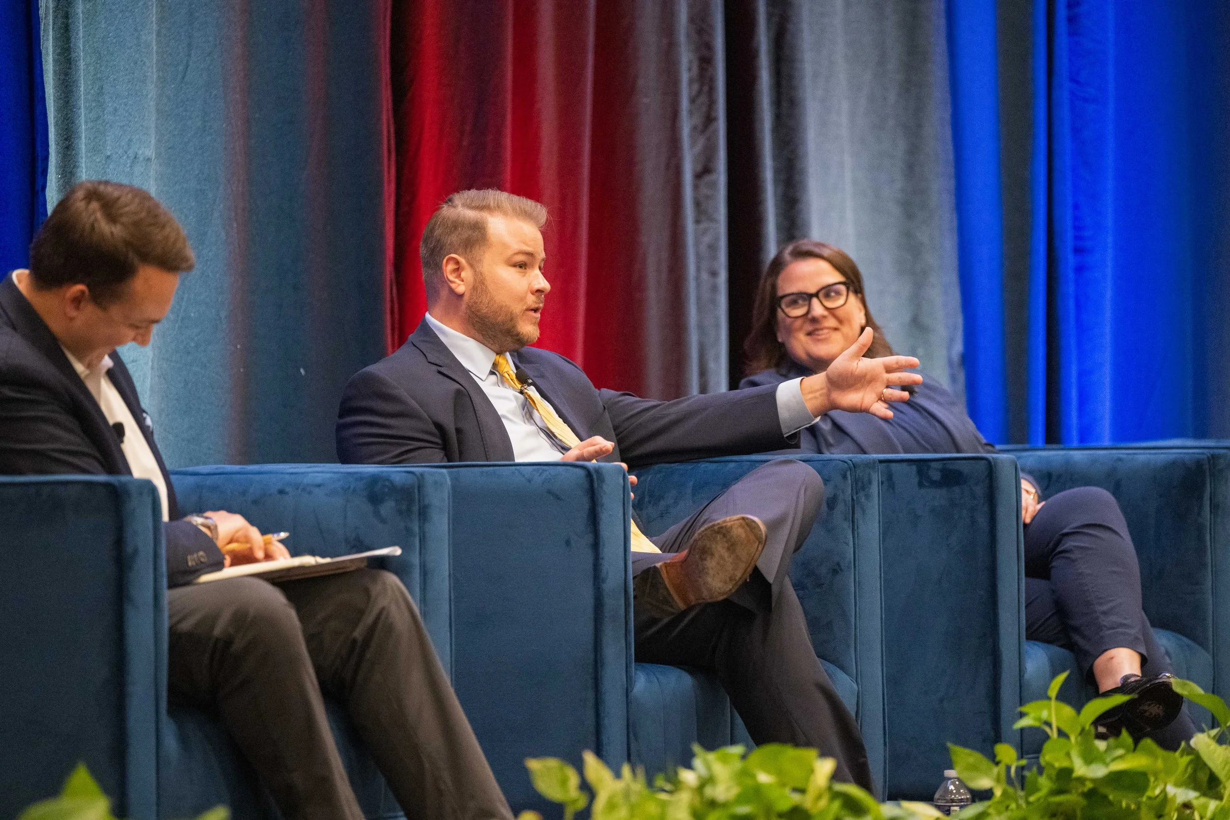 Three people sitting on a blue couch with a colorful curtain backdrop, engaged in a panel discussion. The man in the middle is speaking and gesturing with his hand, while the other two are listening.