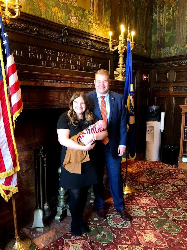 A woman holding a newborn baby, standing next to a man in a suit, inside a formal room with dark wood paneling, an American flag, a blue flag, and a decorative rug.