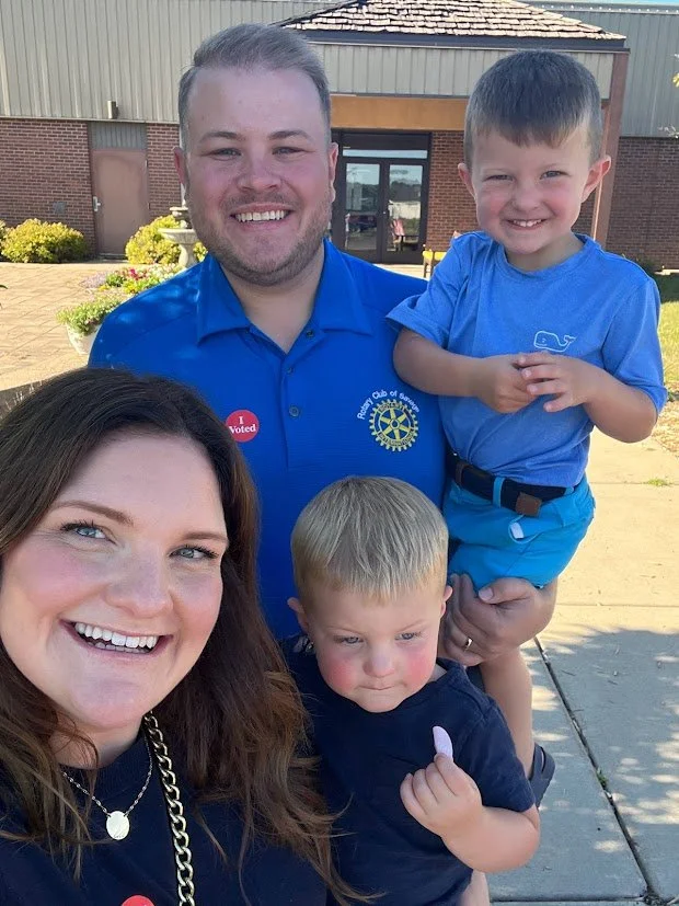 Family group outdoors, smiling together, in front of a building with bushes and potted plants.