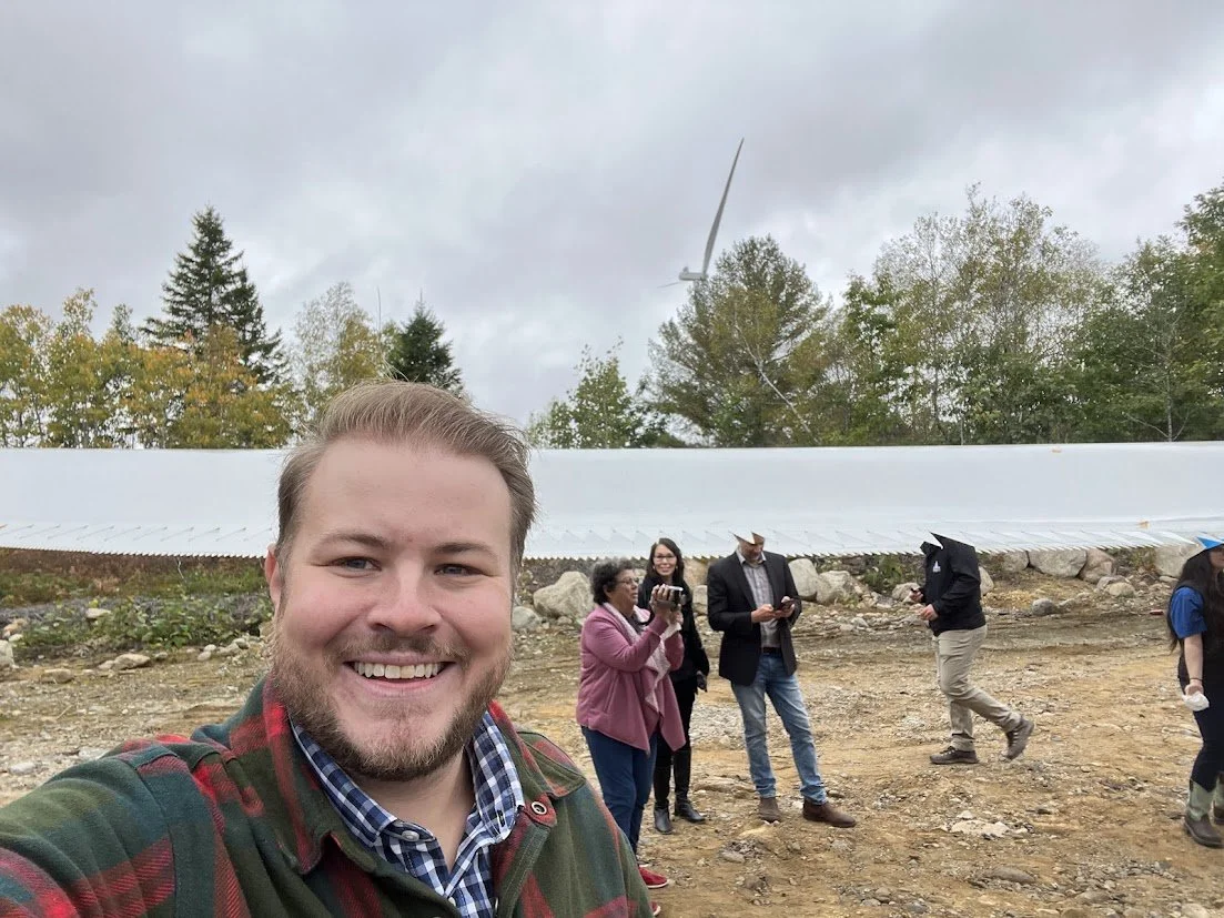 A group of people outdoors, with one man smiling in the foreground taking a selfie. In the background, others are standing and some are taking photos. The setting includes trees and a wind turbine in the distance under a cloudy sky.
