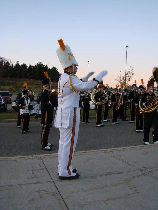 A marching band member in a white uniform with gold trim and a tall white hat with a feather, leading a band of musicians playing brass and percussion instruments outdoors during sunset.