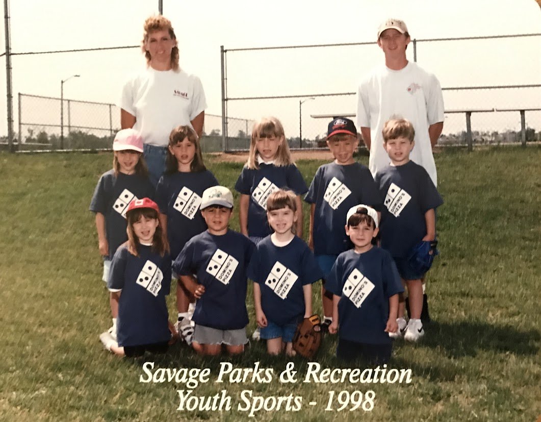 Group of young children and two adults posing on a baseball field in 1998, wearing matching navy blue t-shirts with a domino graphic for Savage Parks & Recreation Youth Sports.