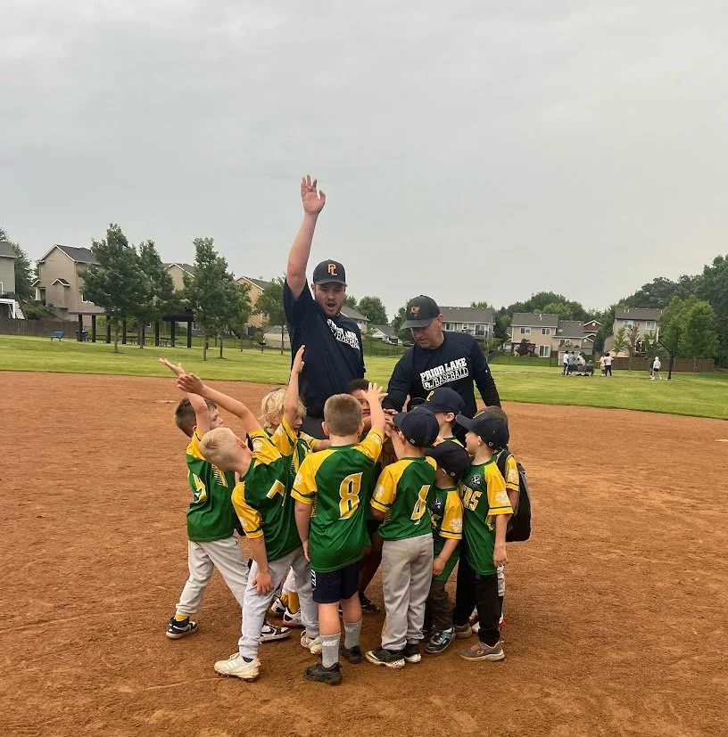 Youth baseball team gathers around coach on field, team wearing green and yellow jerseys, coach raising hand.
