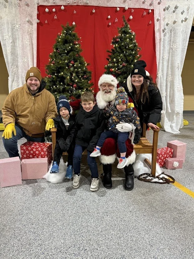 Family sitting with Santa Claus in front of Christmas trees and holiday decorations, with children and adults smiling, surrounded by wrapped gifts.