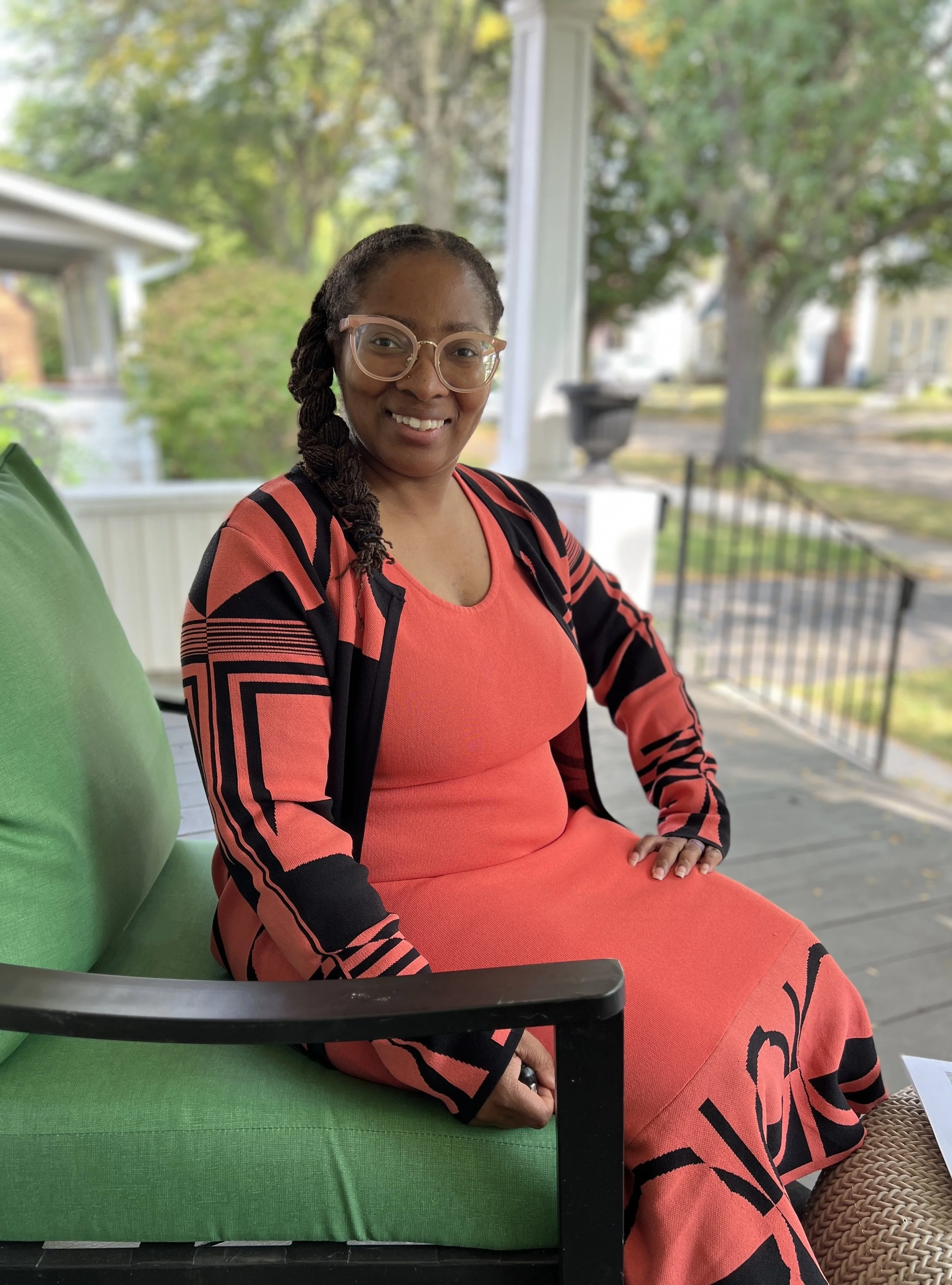 Claretta, a Black woman dressed in coral and black, sits on a porch and smiles at the camera.