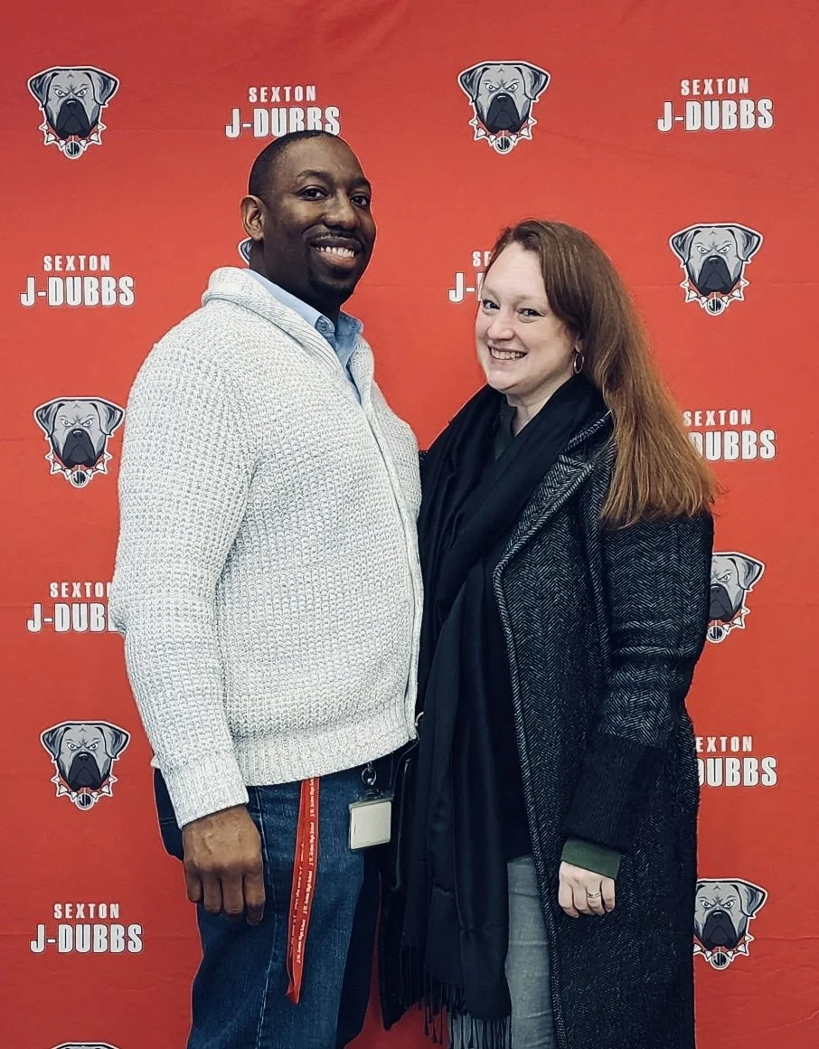 Caitlin and her husband pose at a Sexton H.S. event in front of a step-and-repeat background featuring the Sexton mascot!