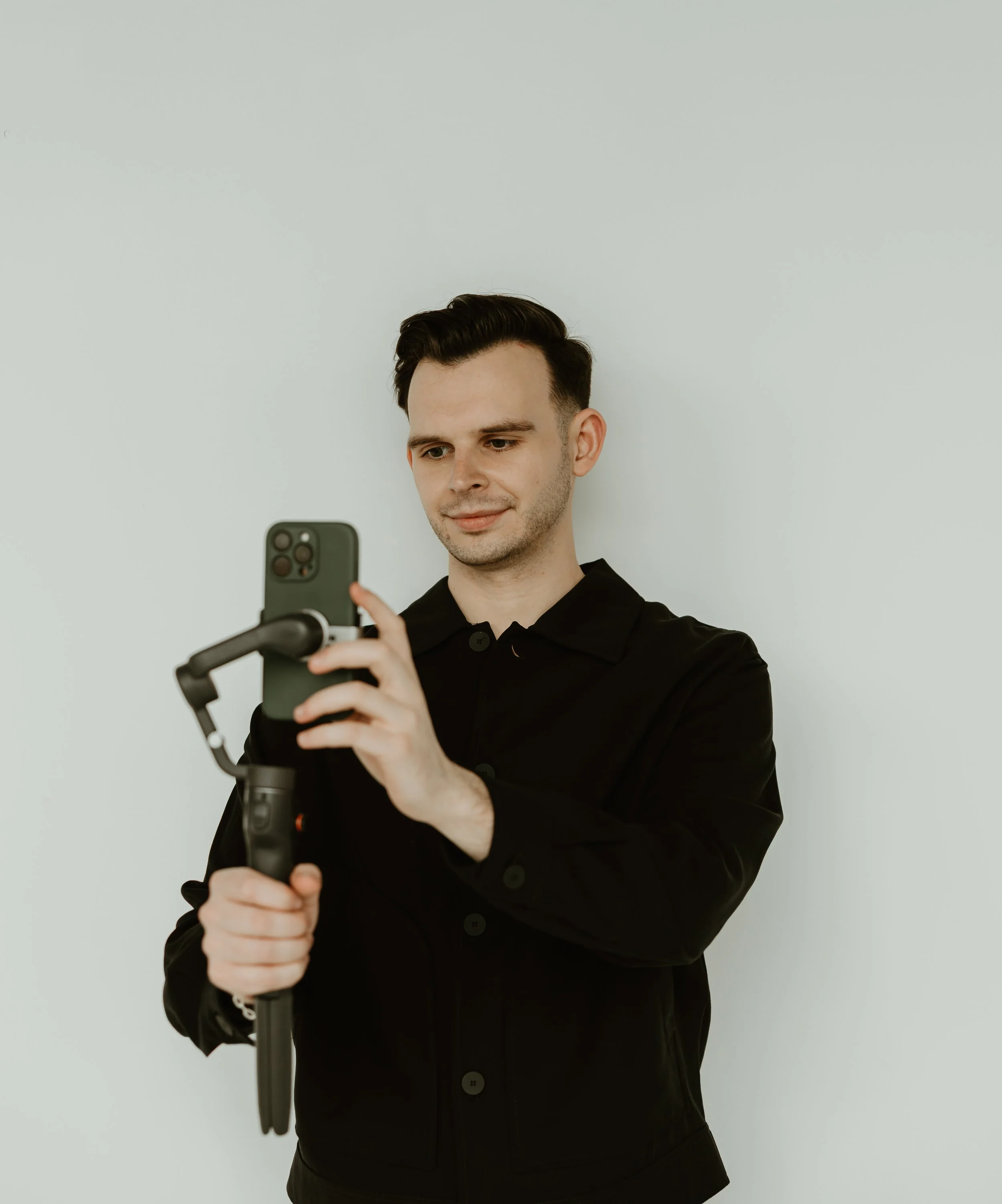 A young man with dark hair, wearing a black jacket, is holding and operating a camera mounted on a handheld stabilizer to take a photo or record a video against a plain white wall background.