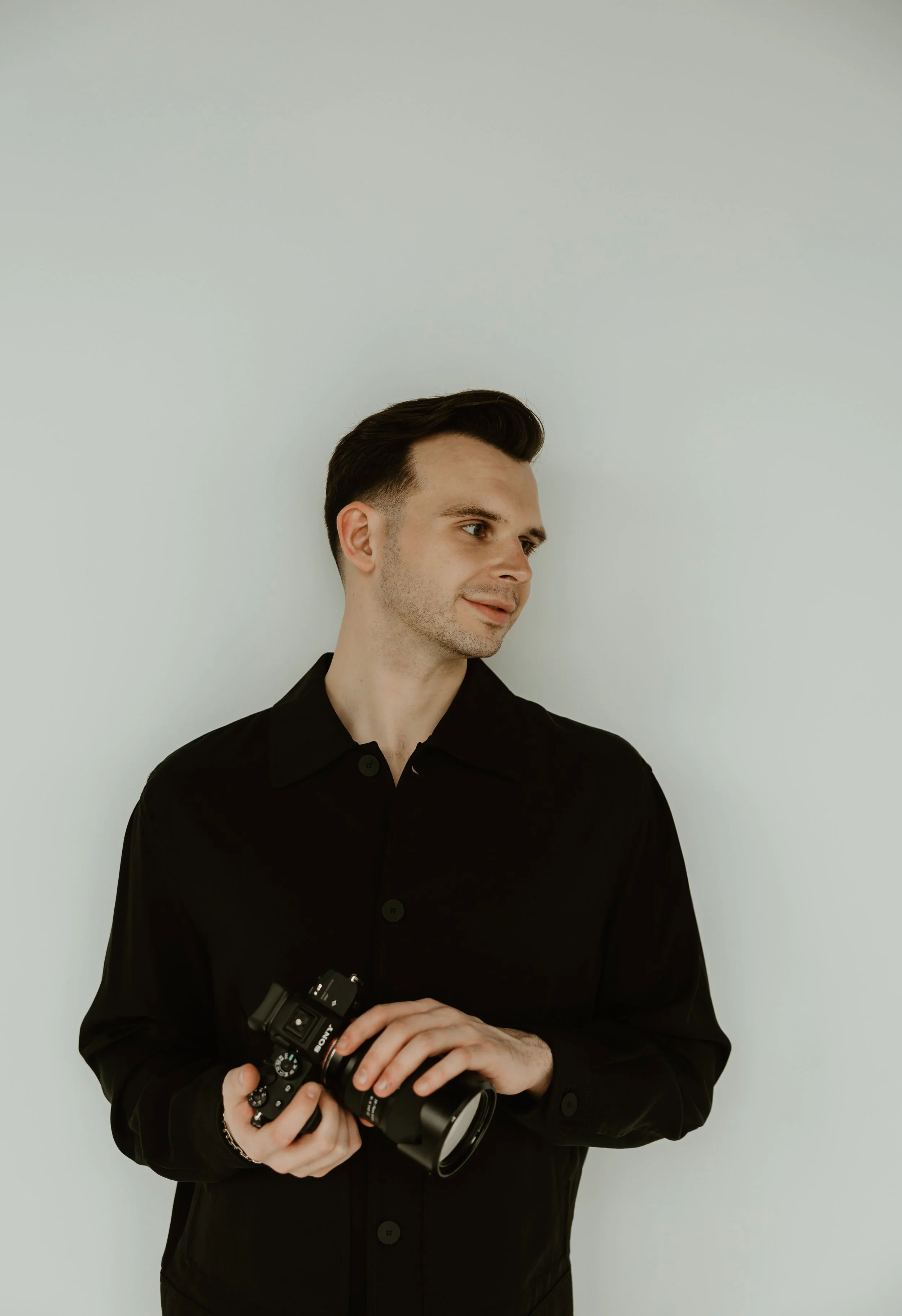 A young man with dark hair, wearing a black shirt, holding a camera in his hands, standing against a plain light-colored background.