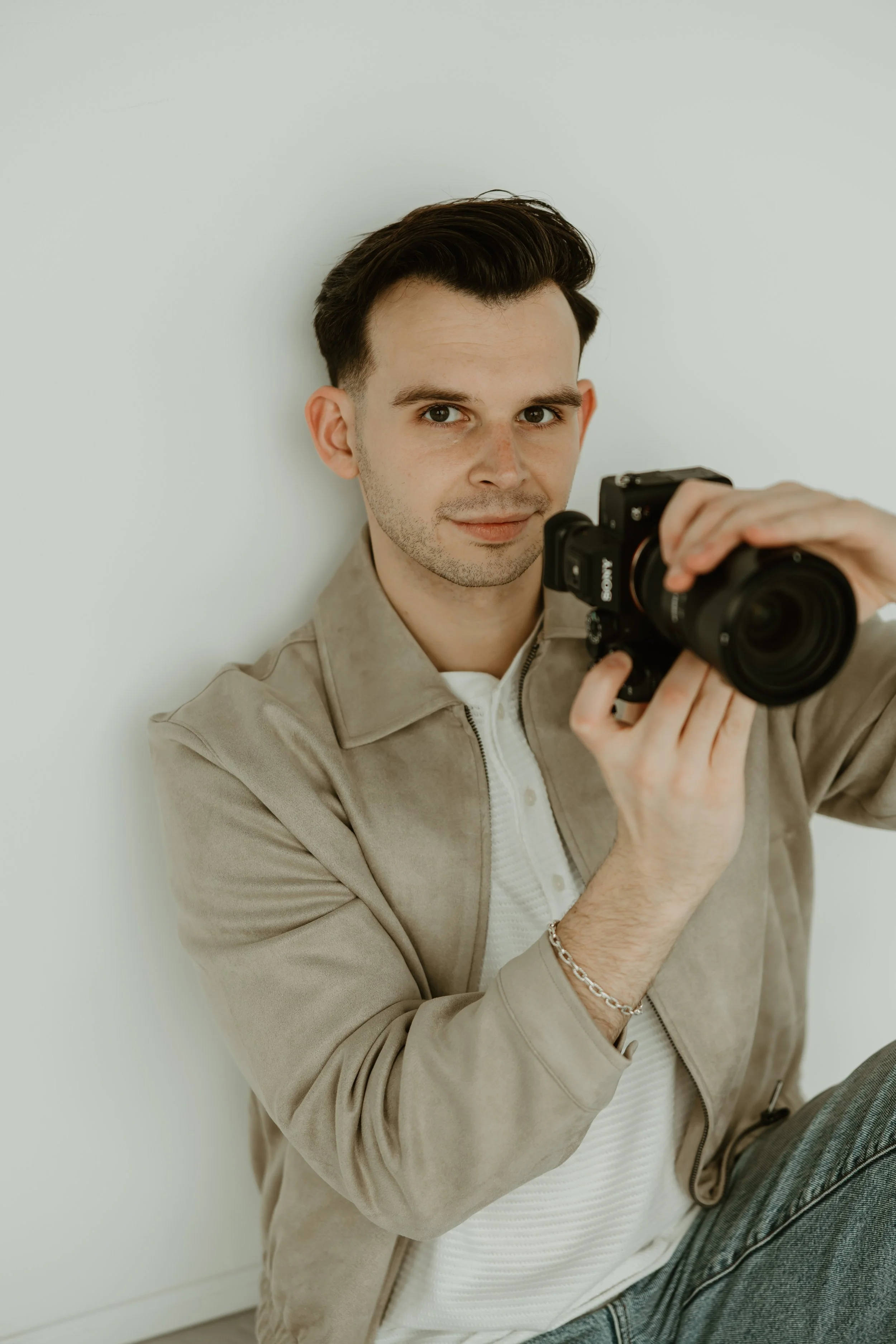 A young man with dark hair, wearing a beige jacket and white shirt, taking a selfie with a professional camera while sitting against a plain white wall.