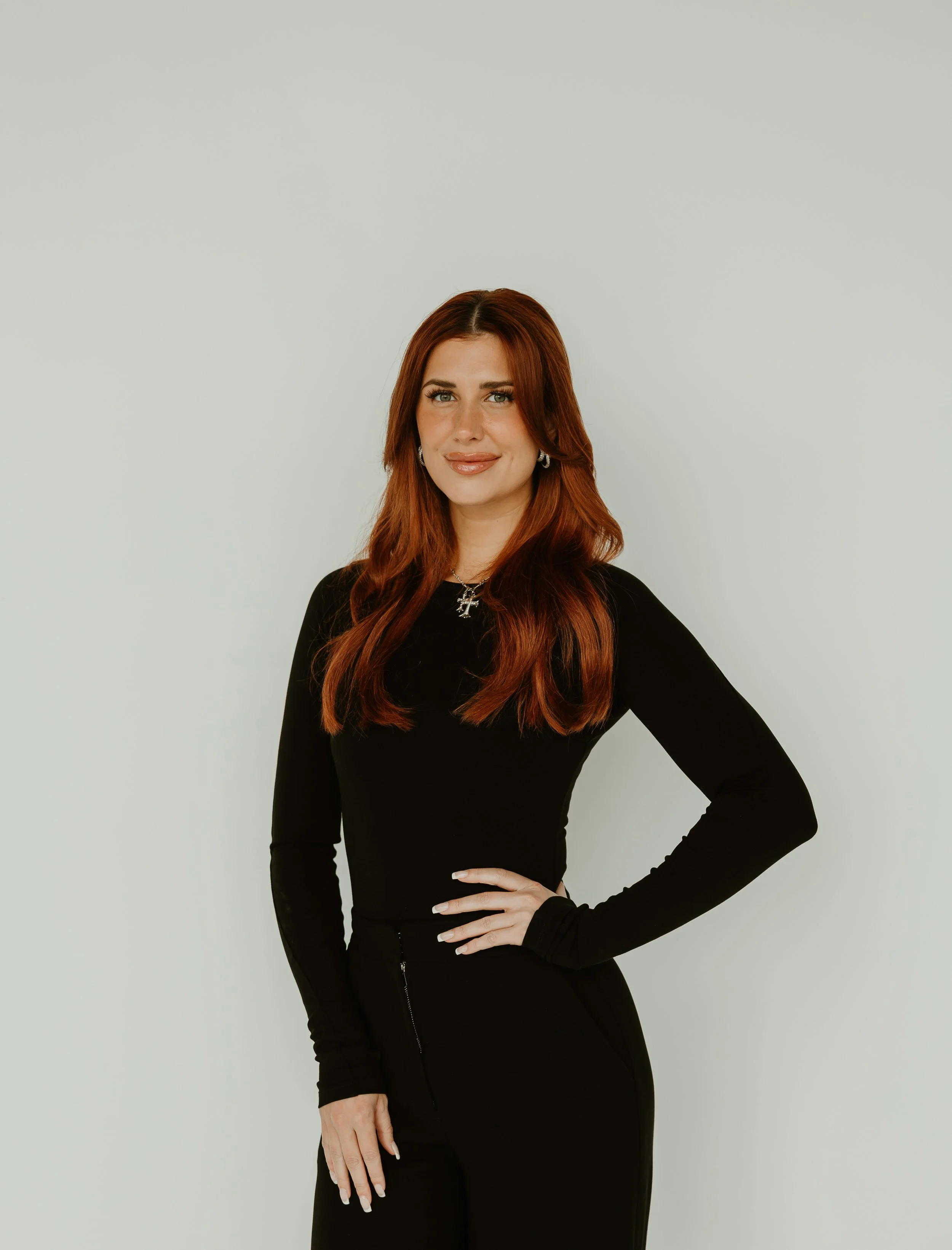 A woman with red hair, wearing a black long-sleeve top and black pants, standing against a plain light-colored background.