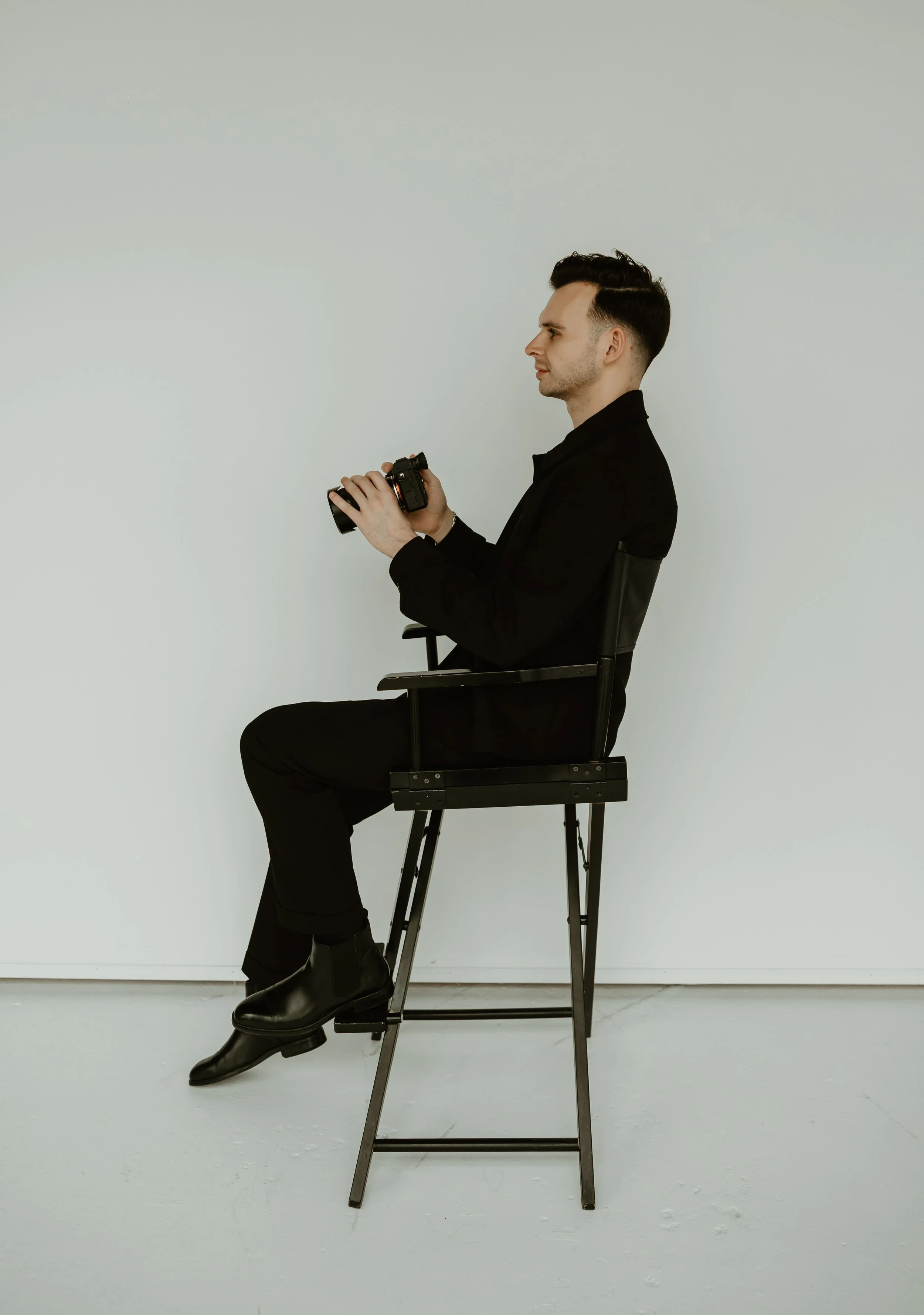 A man dressed in all black sitting on a black chair, holding a camera, against a plain light-colored background.