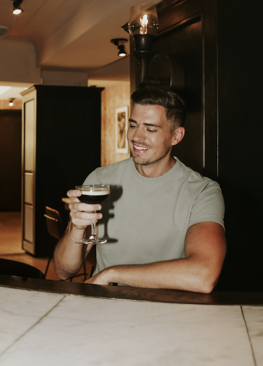 A young man with short dark hair and a light grey t-shirt, smiling and holding a cocktail glass filled with a dark beverage, sitting at a bar in a dimly lit restaurant or bar interior.