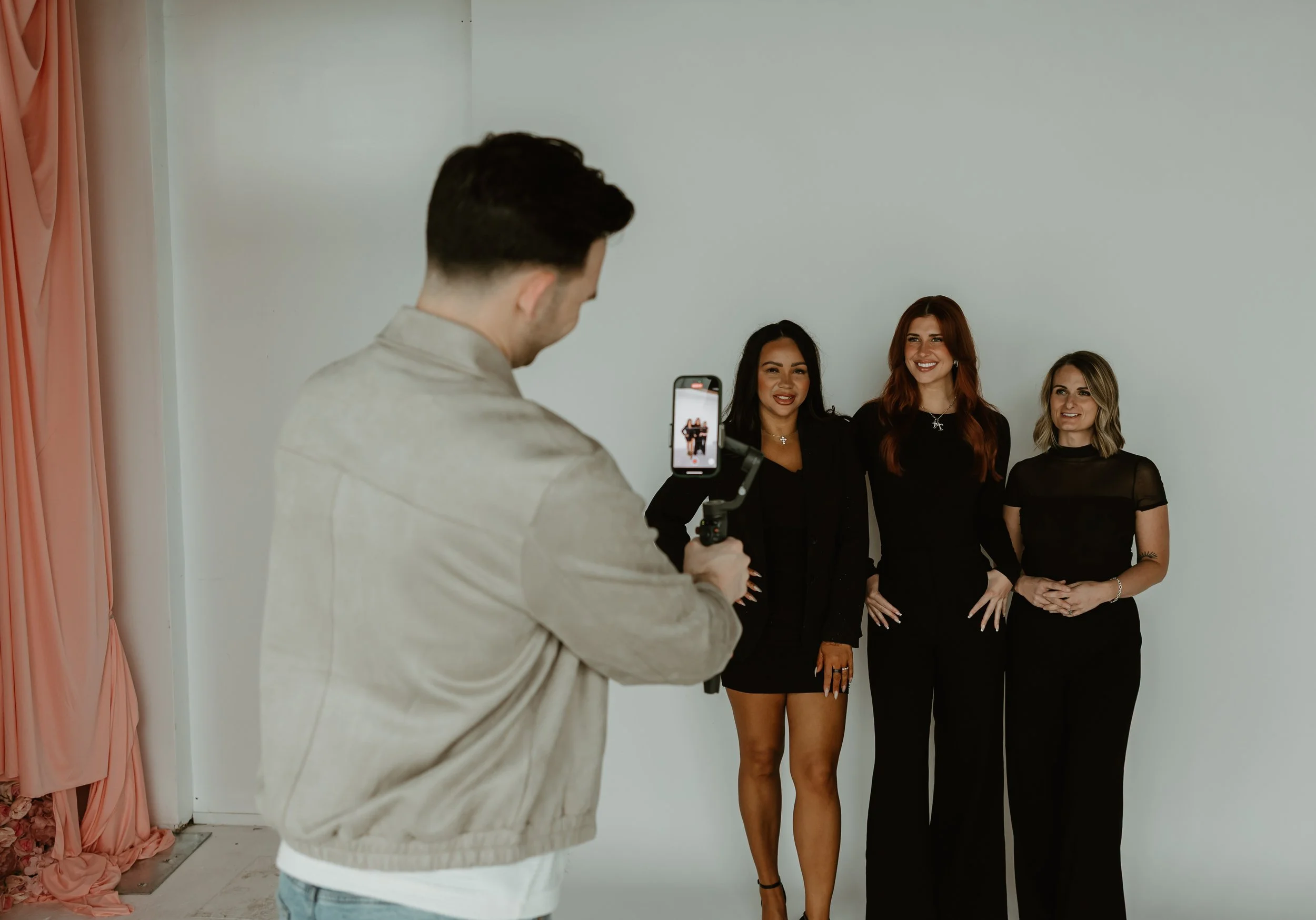 Man taking a photo of three women standing against a white background, with pink curtains on the side.