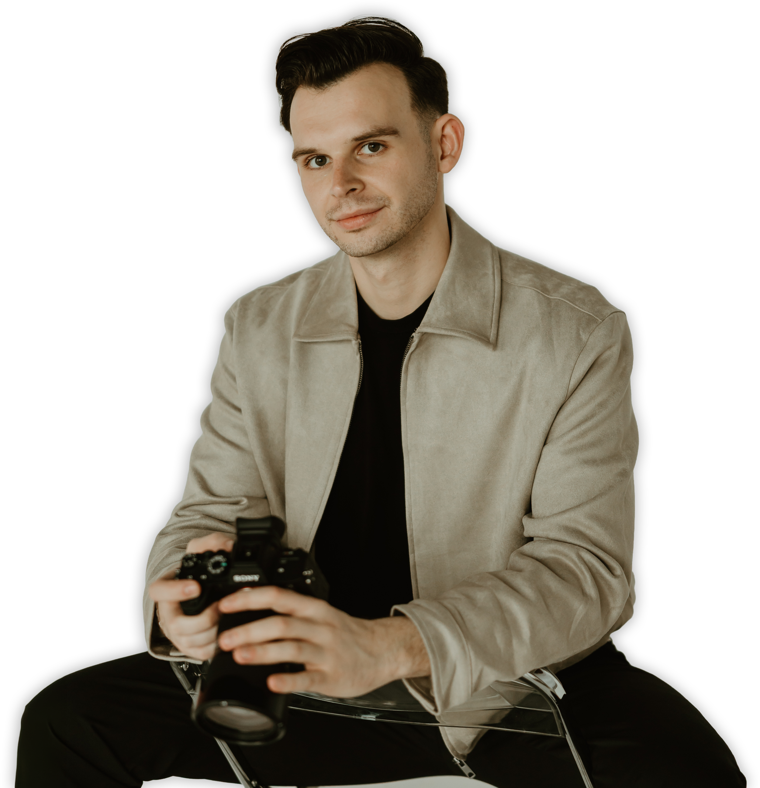 A young man with short dark hair and light skin is sitting on a transparent chair against a black background, holding a Sony camera in his hands. He is wearing a beige jacket over a black shirt and is looking at the camera with a slight smile.