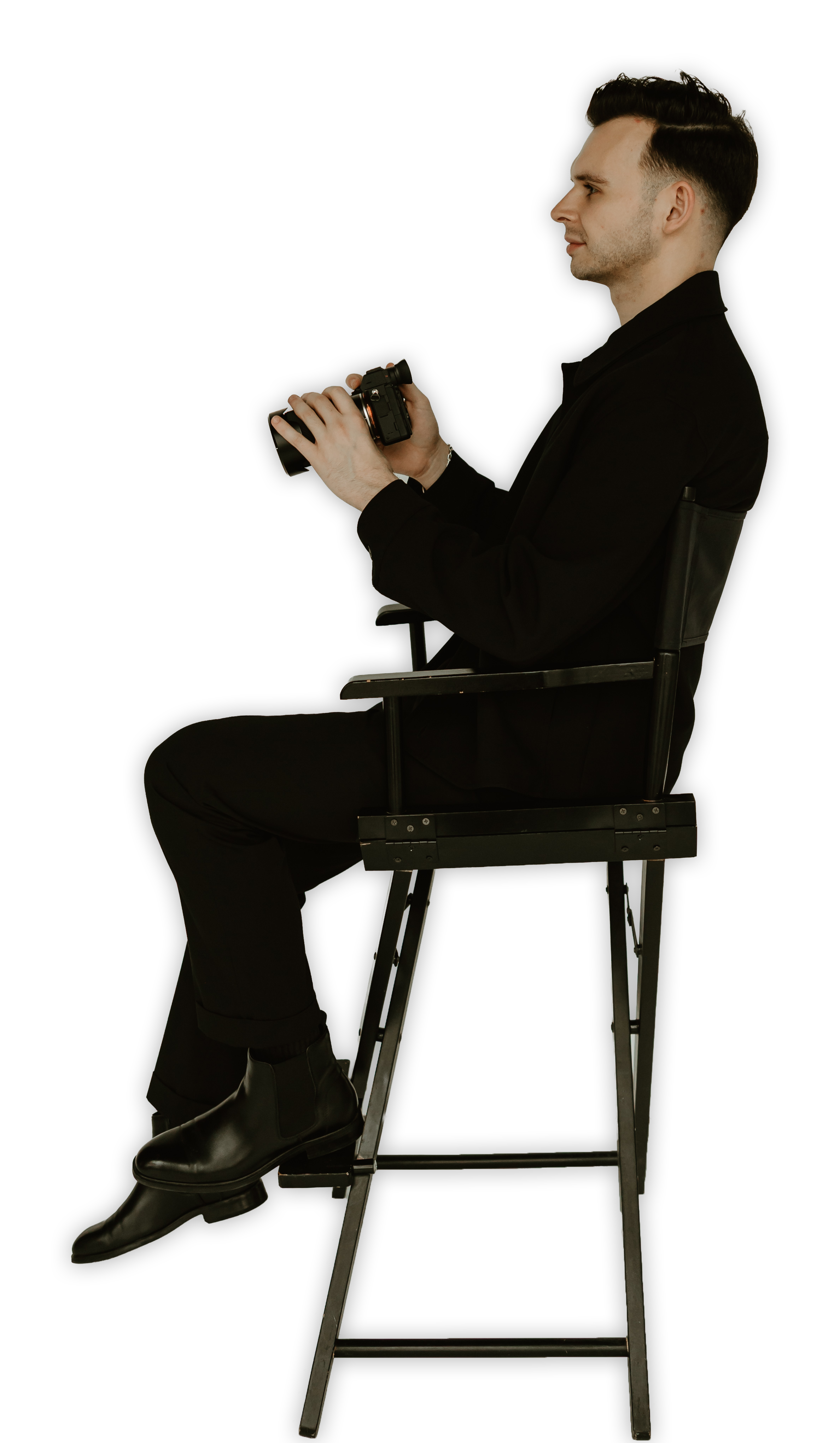 A man dressed in black sitting on a director's chair, holding a camera, against a black background.