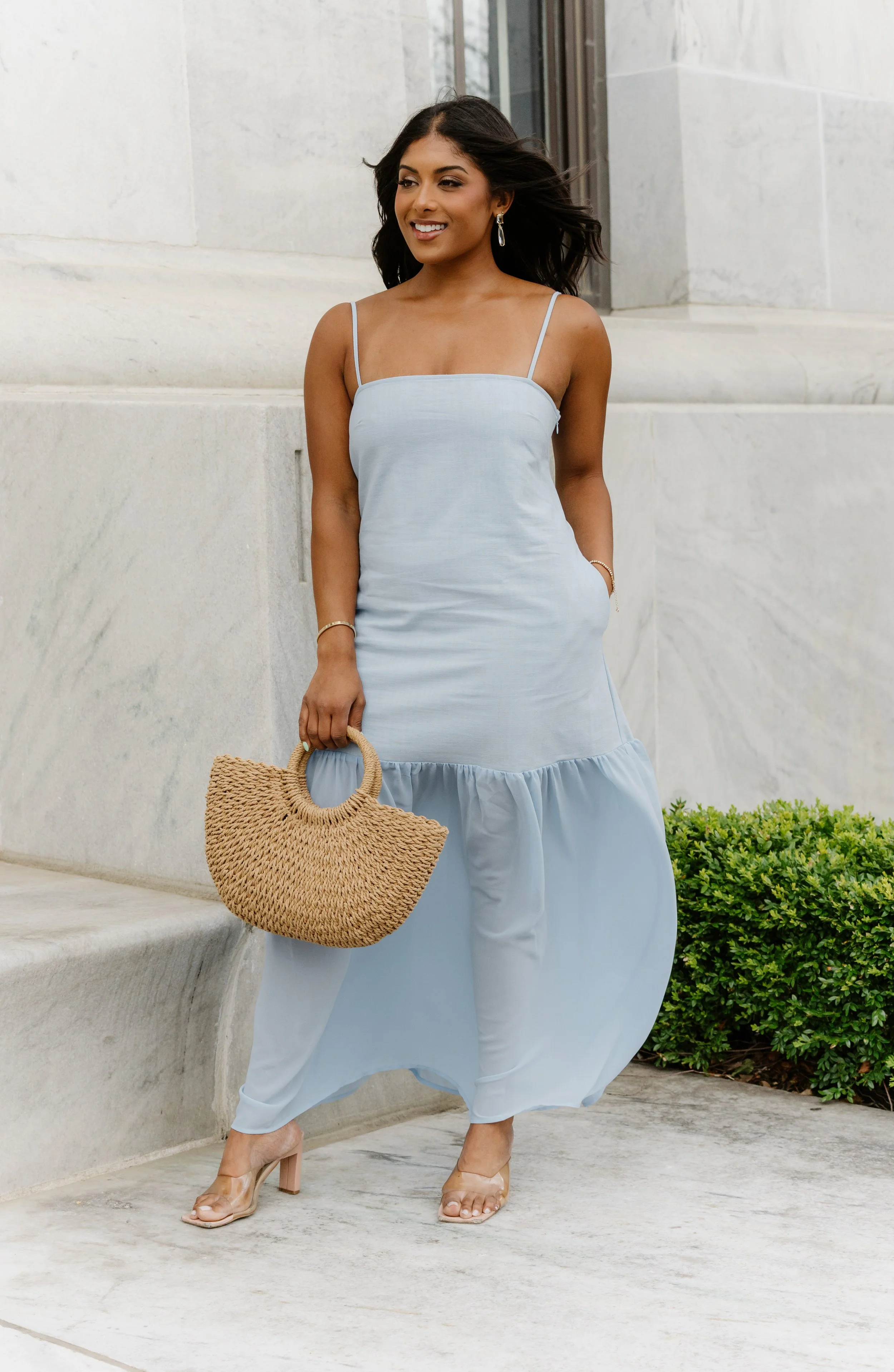 A woman in a light blue sundress holding a woven straw handbag, standing outdoors near a stone building and greenery.