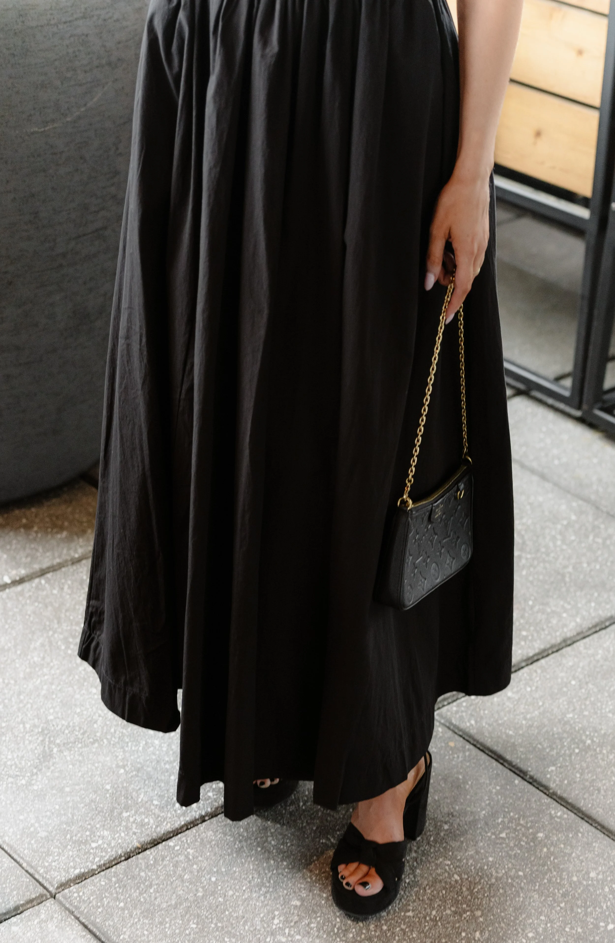 A woman wearing a long black skirt and black shoes, holding a small black purse with a gold chain, standing on a tiled sidewalk.