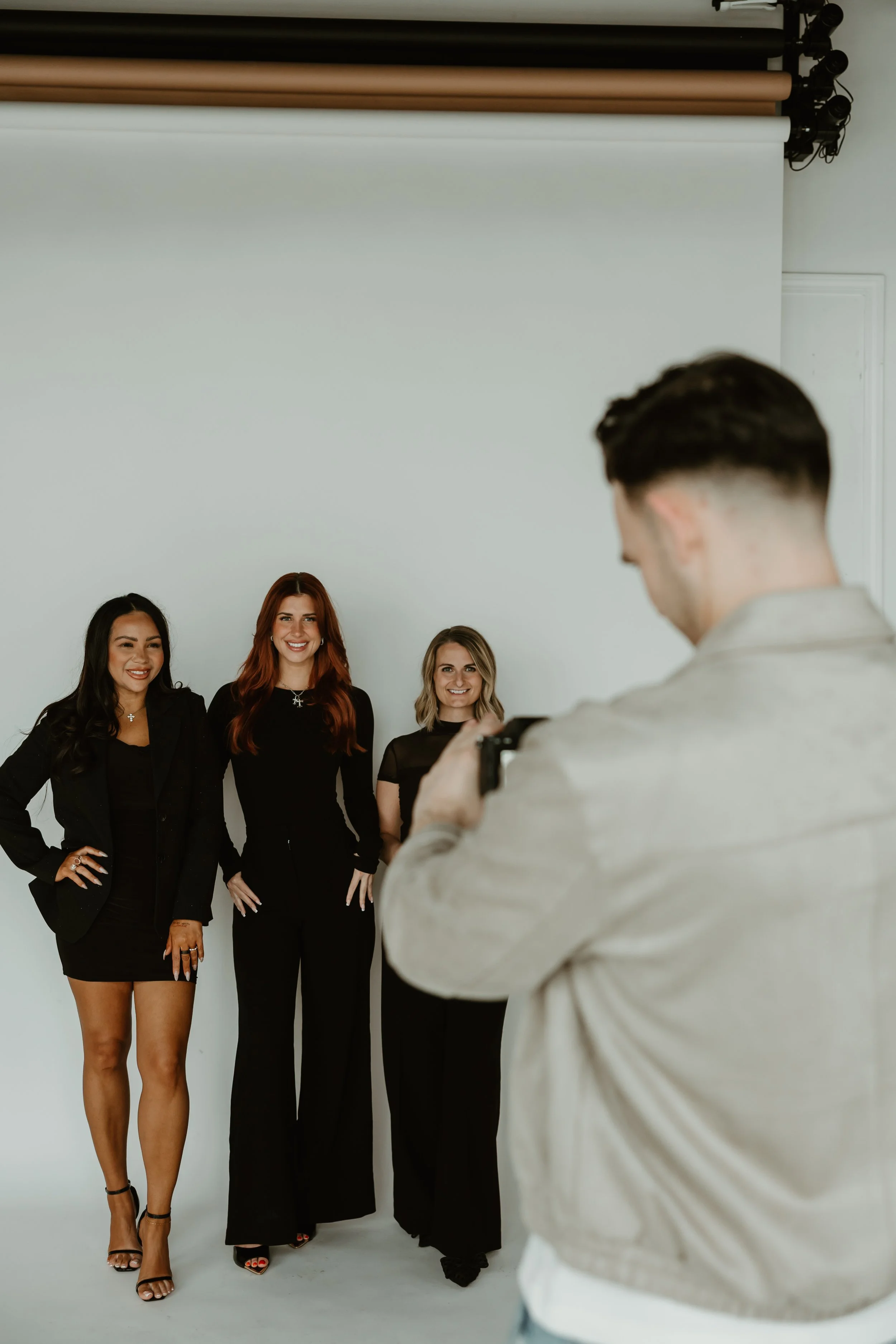 A photographer taking pictures of three women posing in front of a white backdrop in a photo studio.
