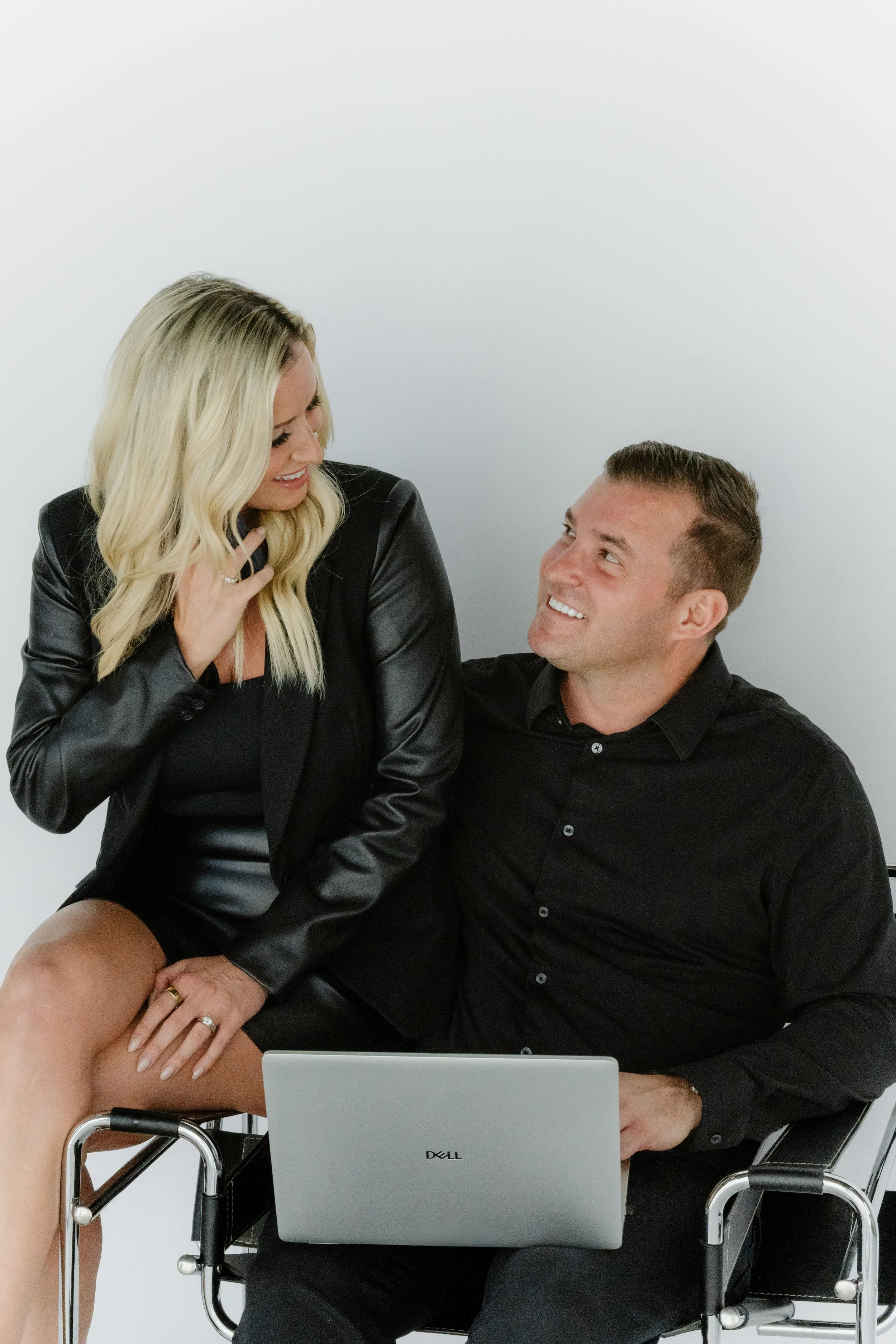 A woman and a man in black clothing share a moment, with the woman sitting on the man's lap while he holds her waist. They are smiling and looking at each other, with a laptop in front of them.