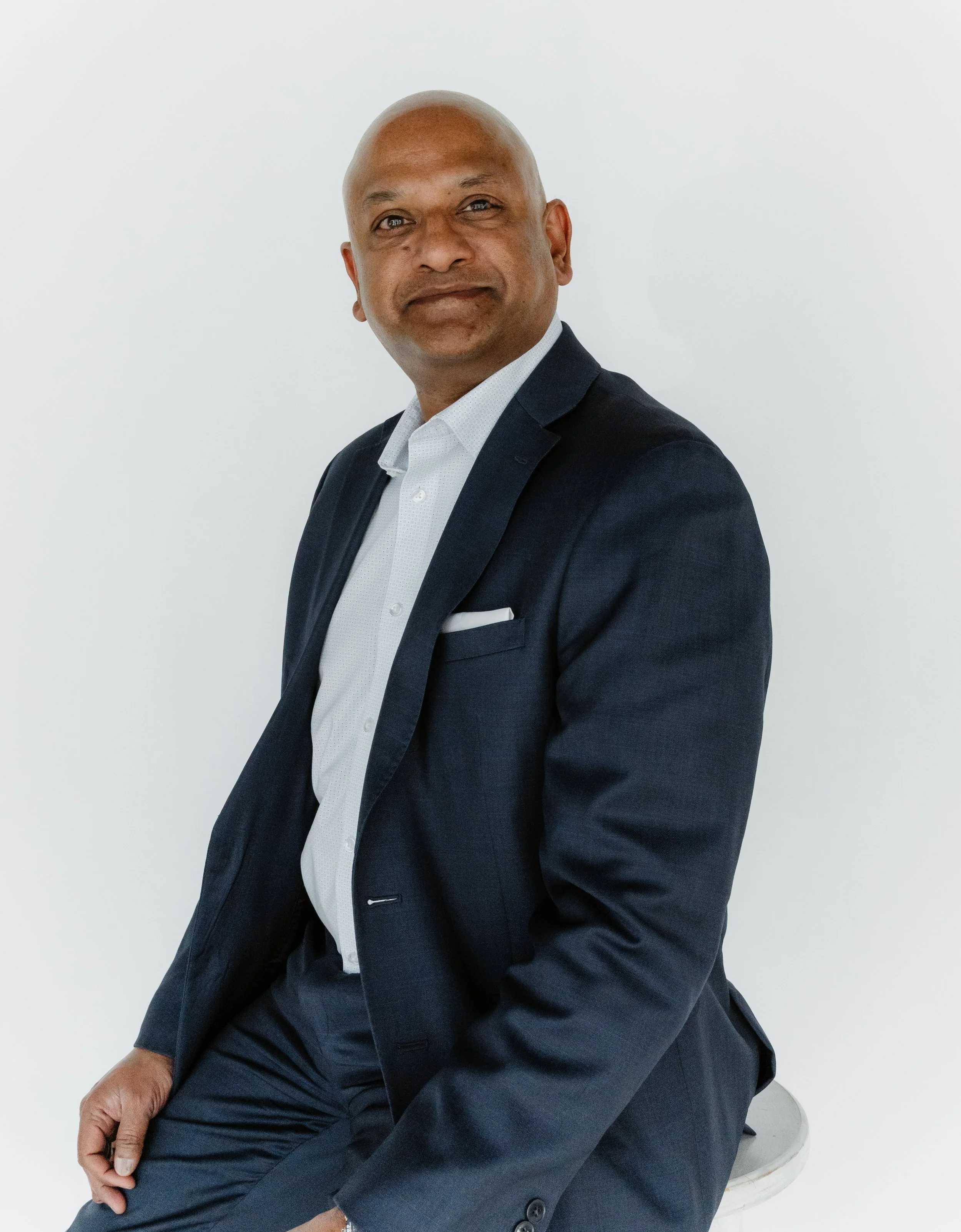 A bald man wearing a dark suit and white shirt sitting on a stool in front of a plain white background.