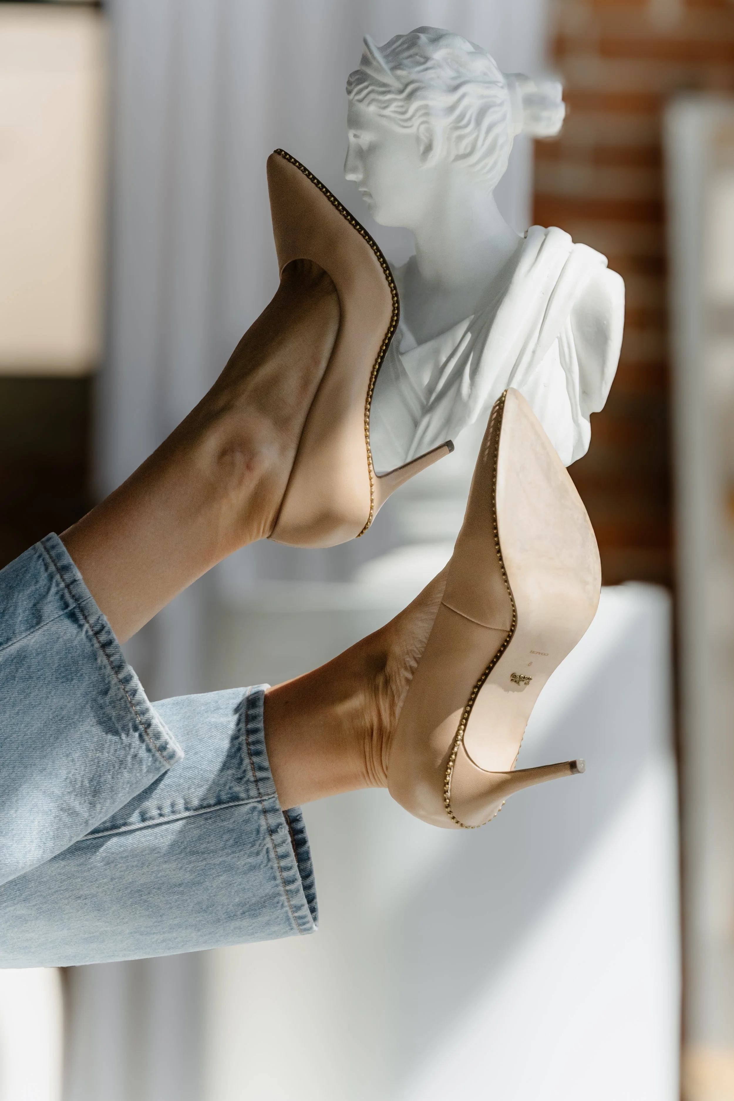 Person holding beige high heel shoes in front of a classical bust sculpture, with a blurred background.