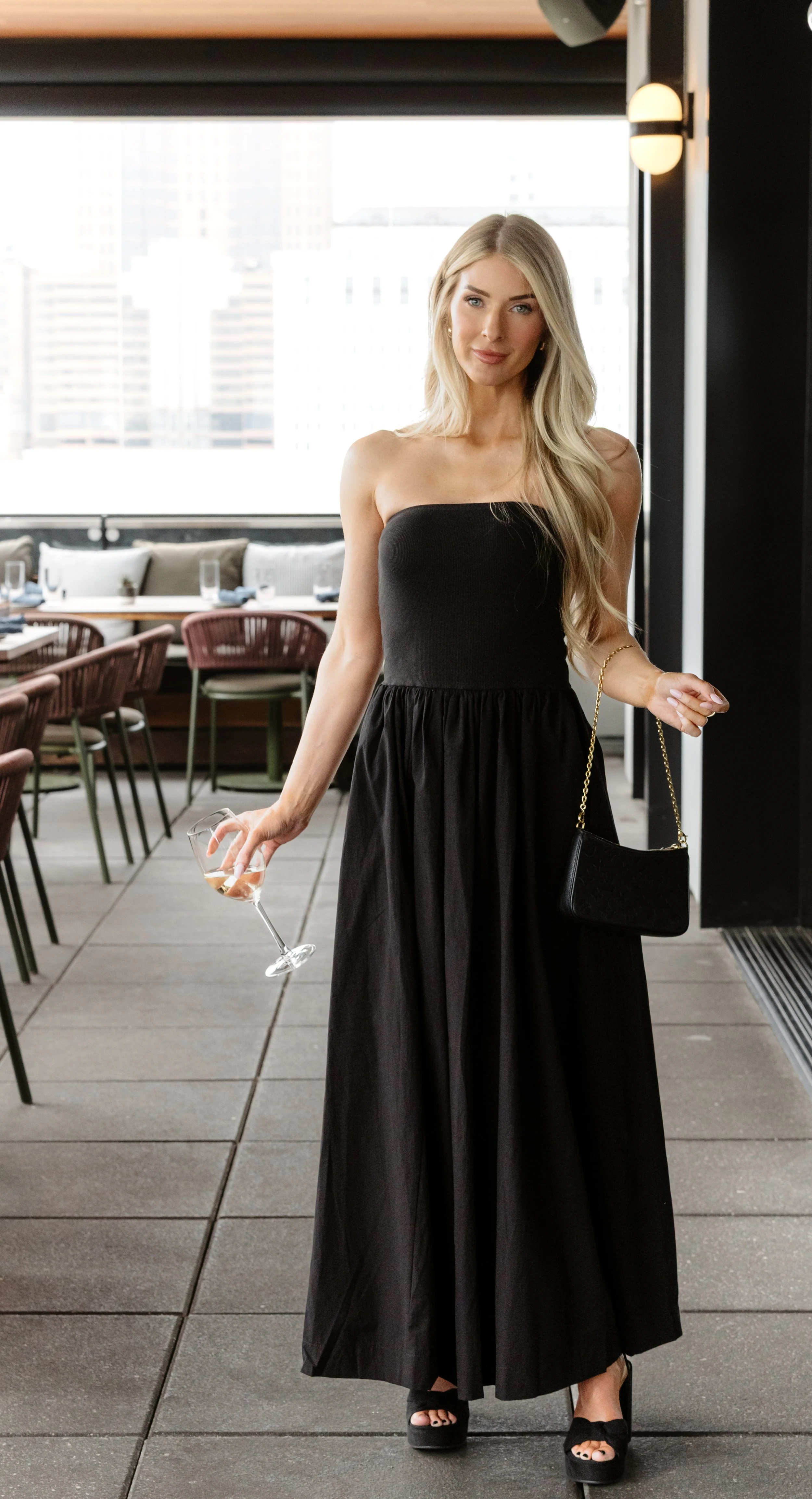 A woman in a strapless black dress holding a glass of white wine standing in a modern indoor restaurant or lounge with large windows and city skyline in the background.