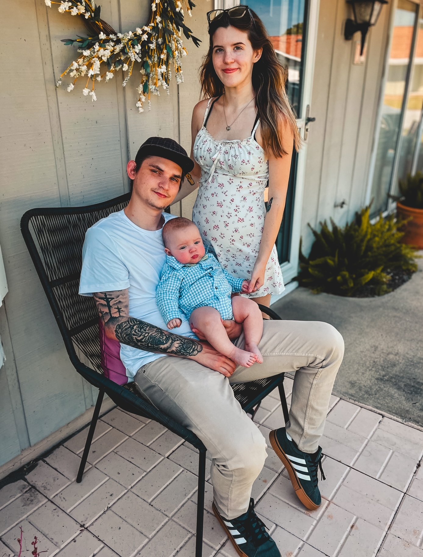 A young woman standing next to a man sitting outdoors on a patio, holding a baby. The woman has long, wavy brown hair, glasses on her head, and is wearing a white dress with small floral prints. The man has short dark hair, tattoos on his arms, and is wearing a white t-shirt, beige pants, and a black cap. The baby is dressed in a blue checkered shirt and shorts, with a curious expression. They are in front of a light-colored wall decorated with a white flower wreath, and there are plants in the background.