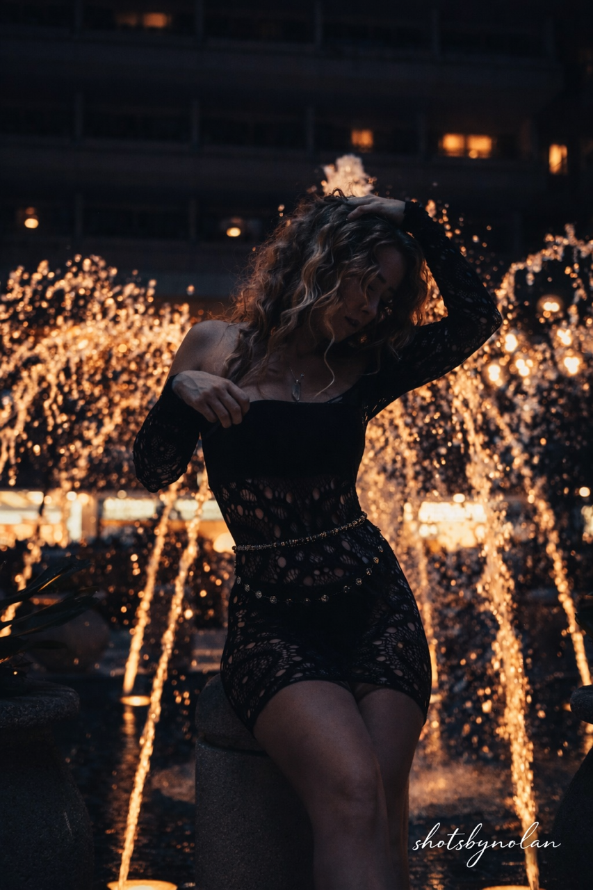 A woman in a black lace dress posing in front of a water fountain with illuminated sparks during sunset.