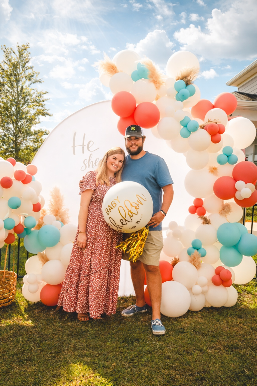 A smiling couple standing together outdoors during daytime, celebrating a pregnancy reveal with balloons and an arch made of white, coral, and turquoise balloons, and a round sign that reads 'Baby theolan is,