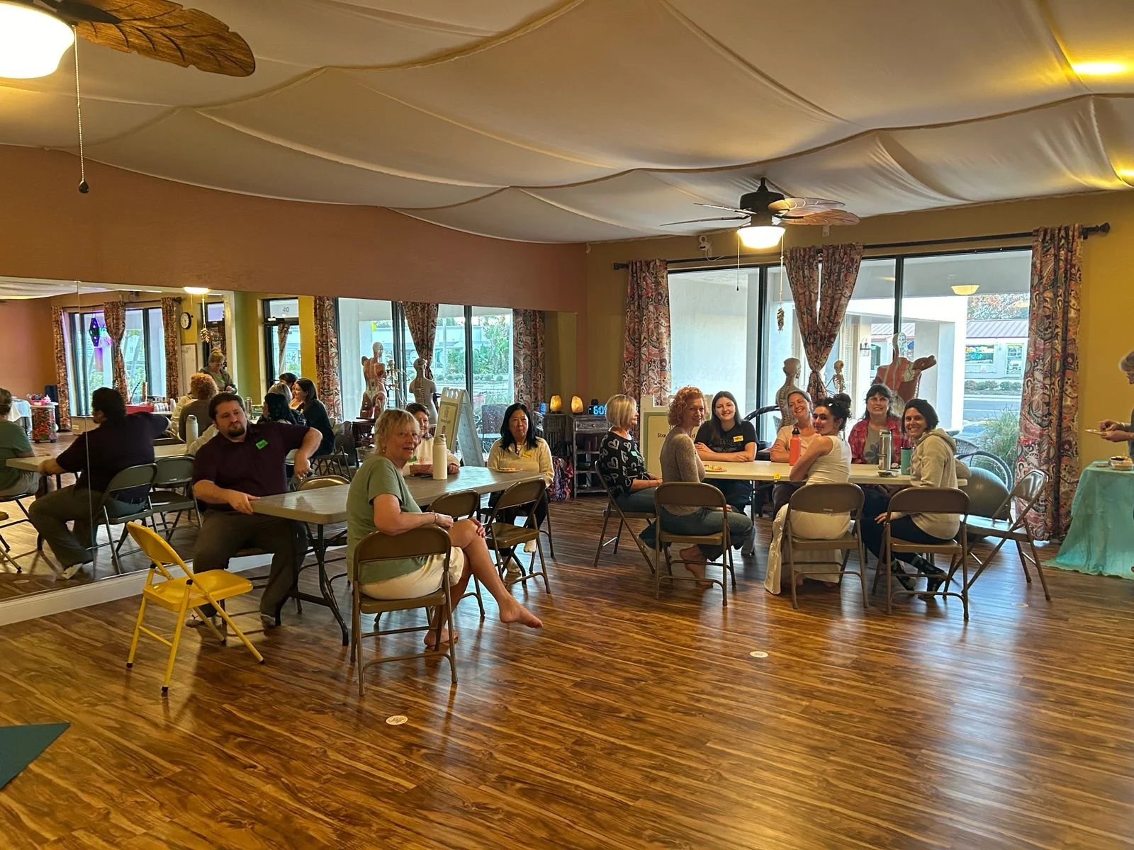 A group of people sitting around tables in a room with large windows and floral curtains, smiling and engaging with each other.