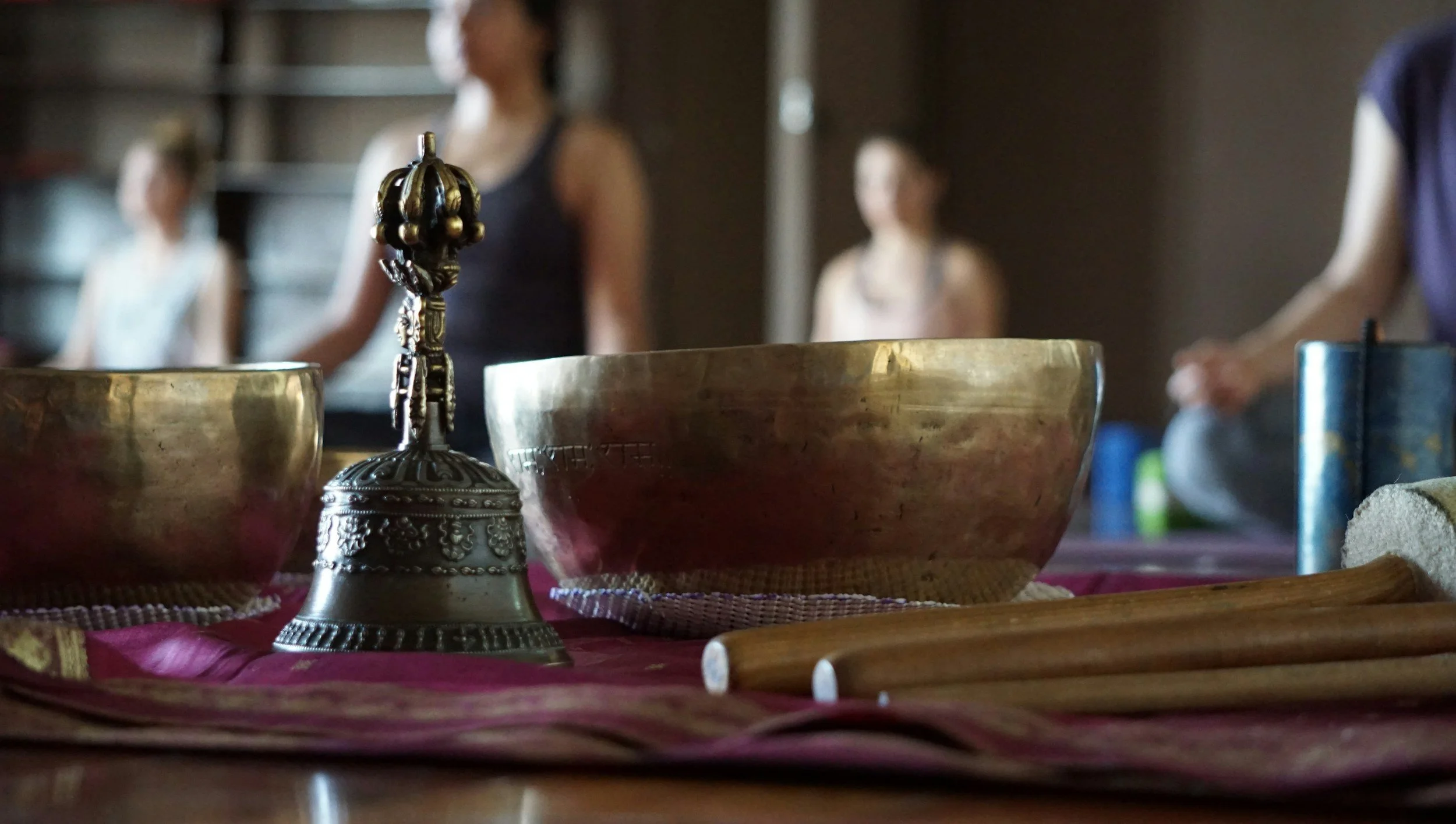 Close-up of brass bowls and a small decorative bell on a table with a pink cloth, with people standing in the background.