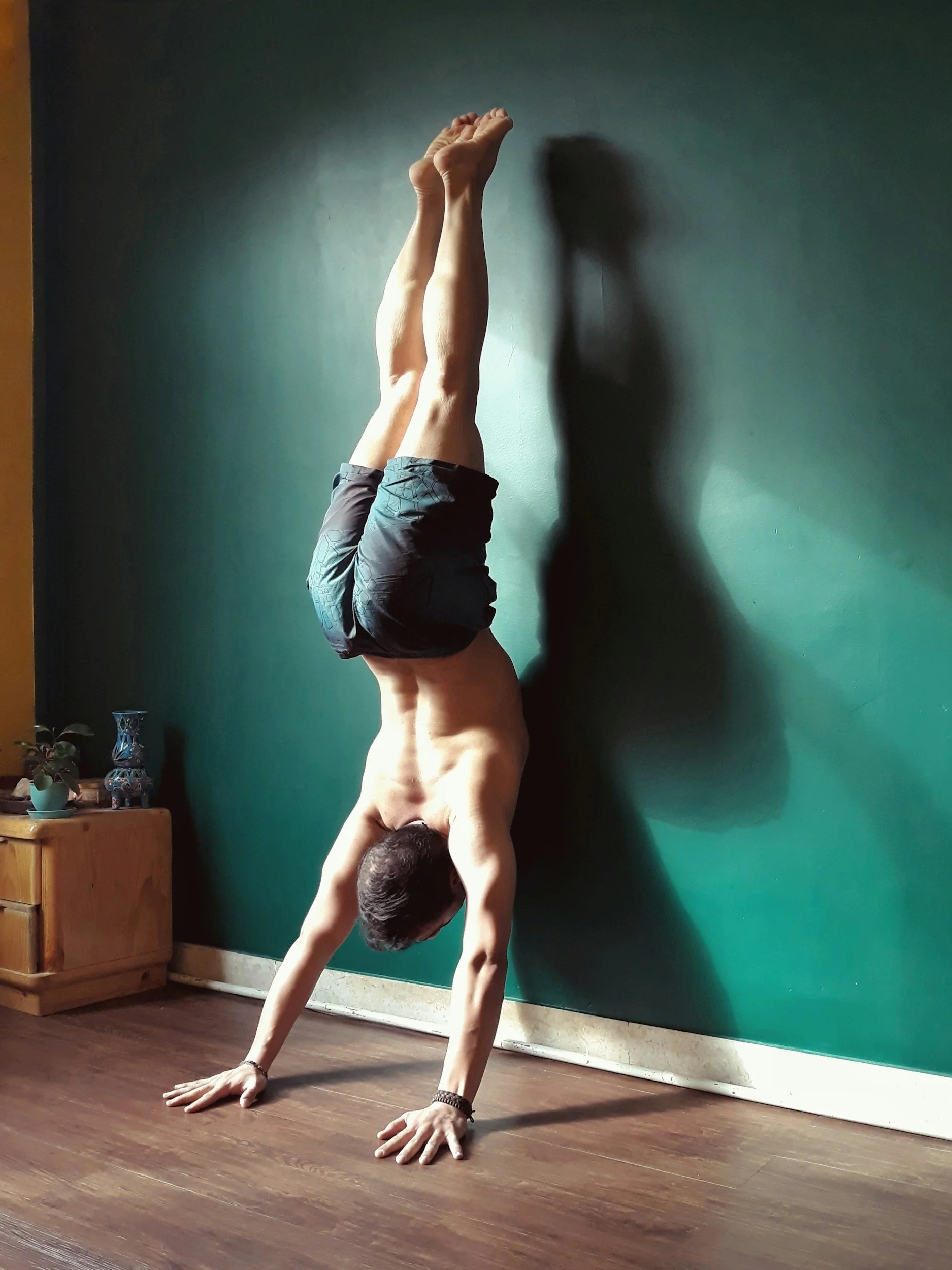 A man performing a handstand against a green wall in a room with wooden floors, with his legs straight up and arms supporting his body, wearing black shorts and wristbands.