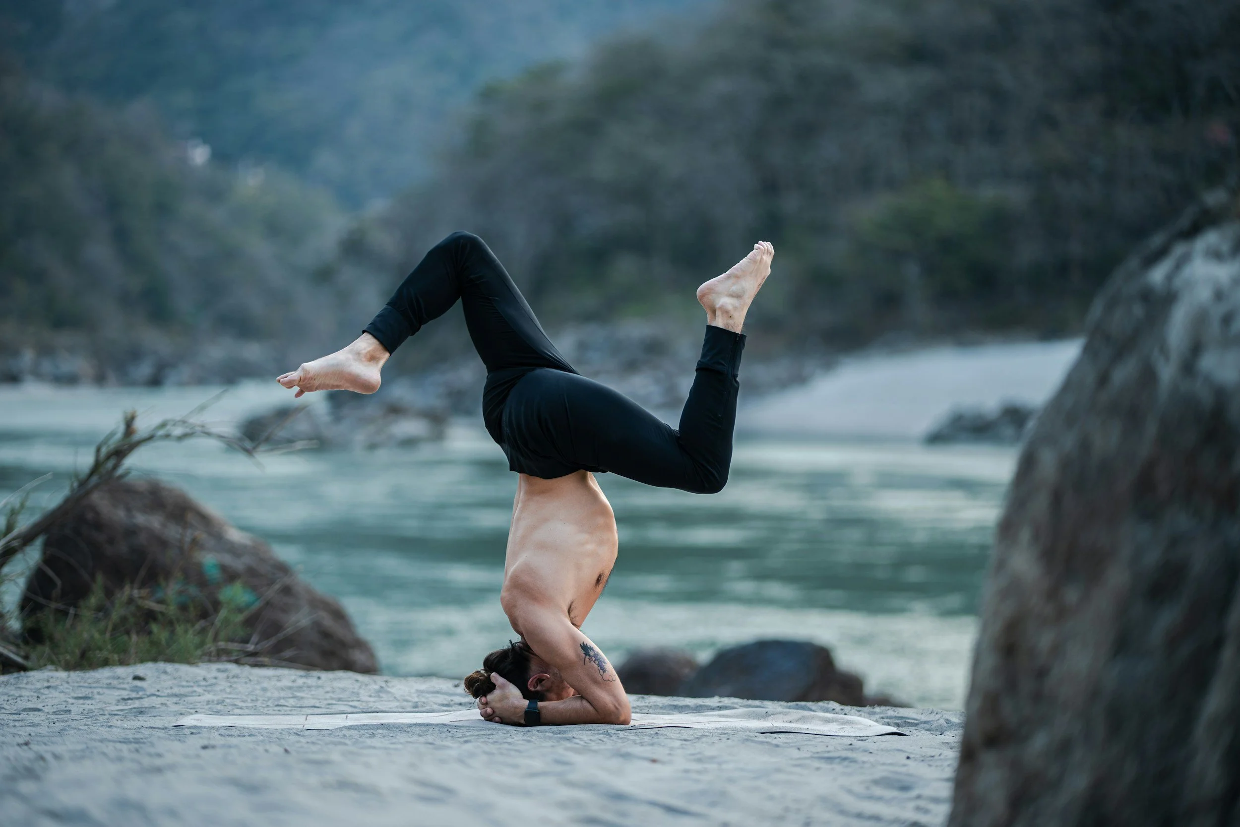 A person practicing yoga in a headstand position on a beach, with their hands clasped behind their head and their legs bent backward, near rocks and the ocean.