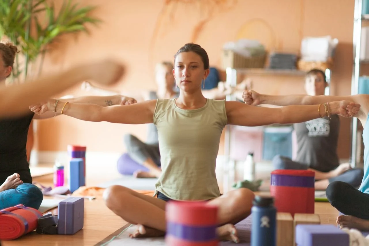 A woman practicing yoga seated cross-legged with arms extended in a yoga studio surrounded by other people and yoga props.