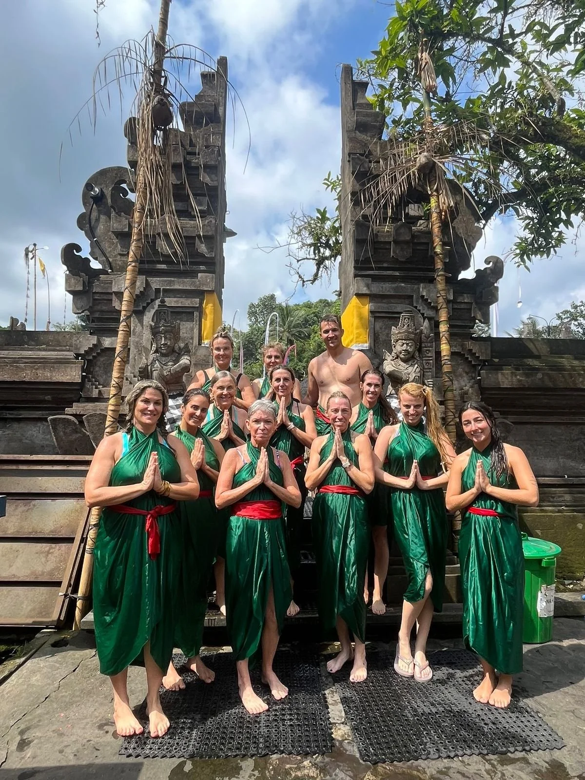 A group of people dressed in green traditional outfits with red sashes, standing barefoot in front of a historical stone gateway, engaging in a prayer or meditation pose with hands in a prayer position. The gateway features carvings and statues, with a partly cloudy sky and greenery in the background.
