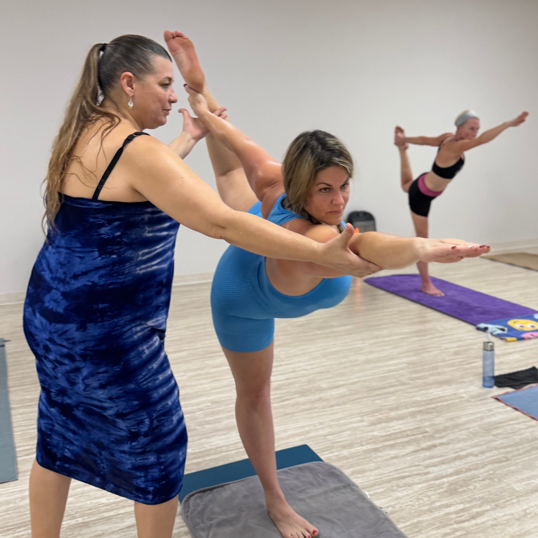 Two women practicing yoga in a studio, with a third woman in the background doing a balancing pose on a purple yoga mat.