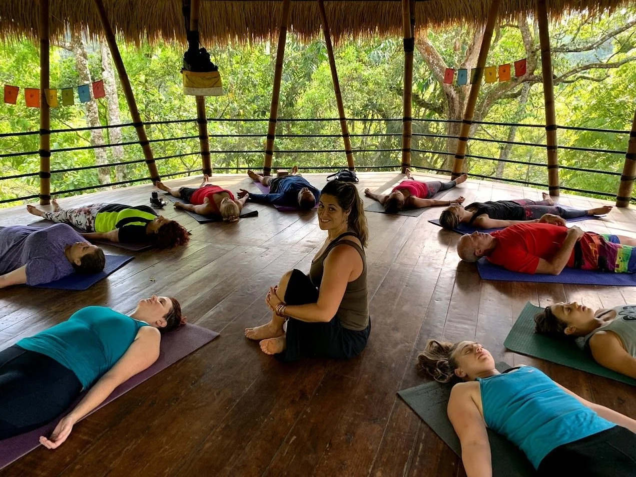 Group of people participating in a yoga class on a wooden deck surrounded by trees, with a yoga instructor sitting on the floor smiling at the camera.