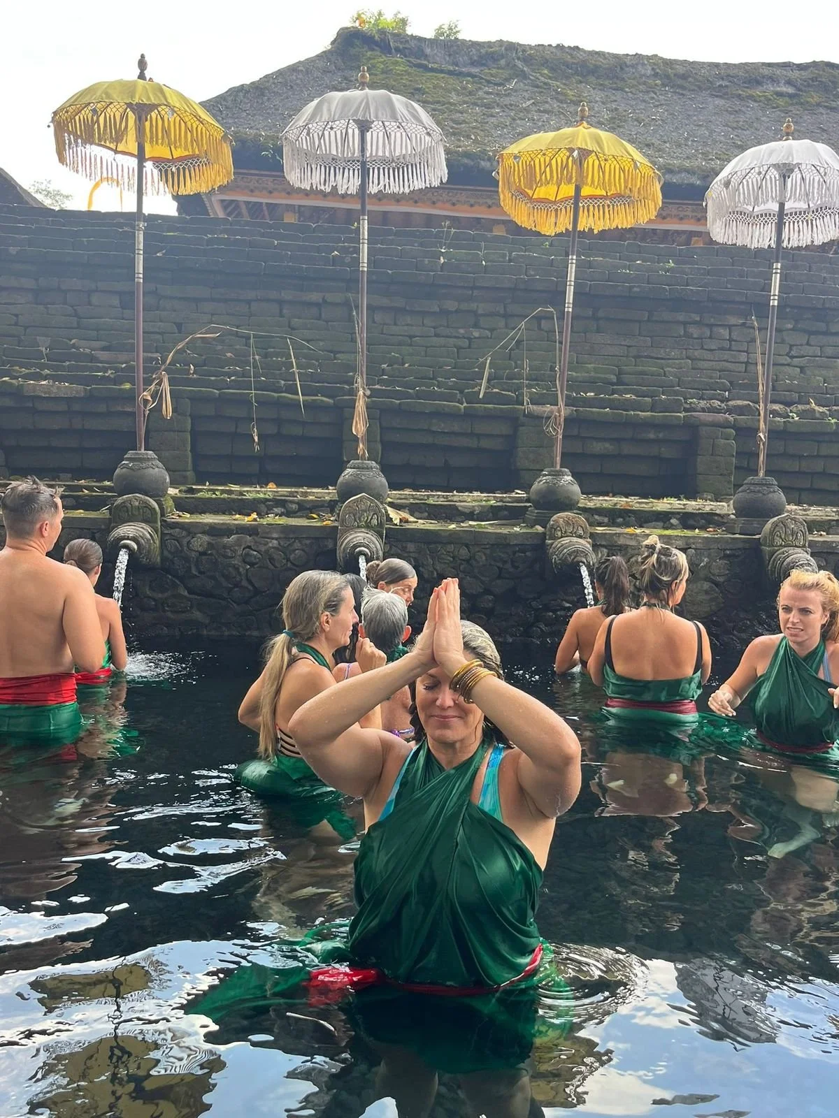 Group of people practicing meditation in a natural hot spring with water spouts and Balinese-style umbrellas in the background.