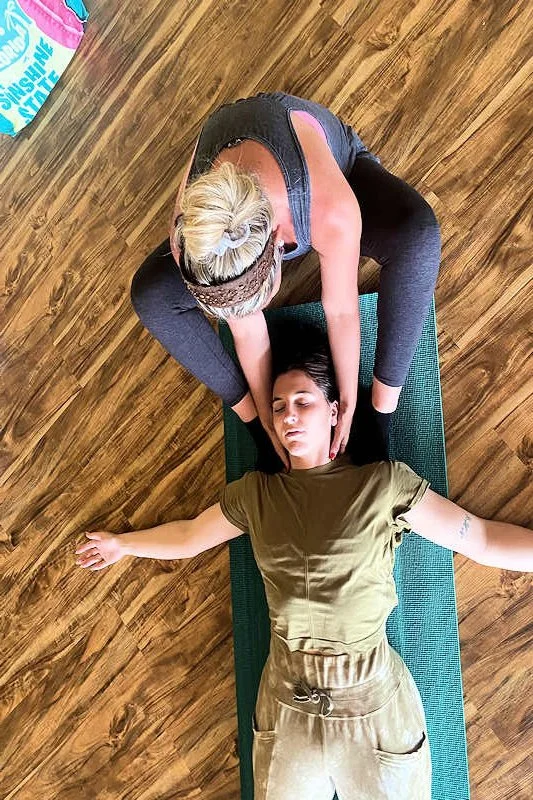 A person with blonde hair practicing yoga or meditation while another person assists with a head or neck massage on a yoga mat in a wooden-floored room.