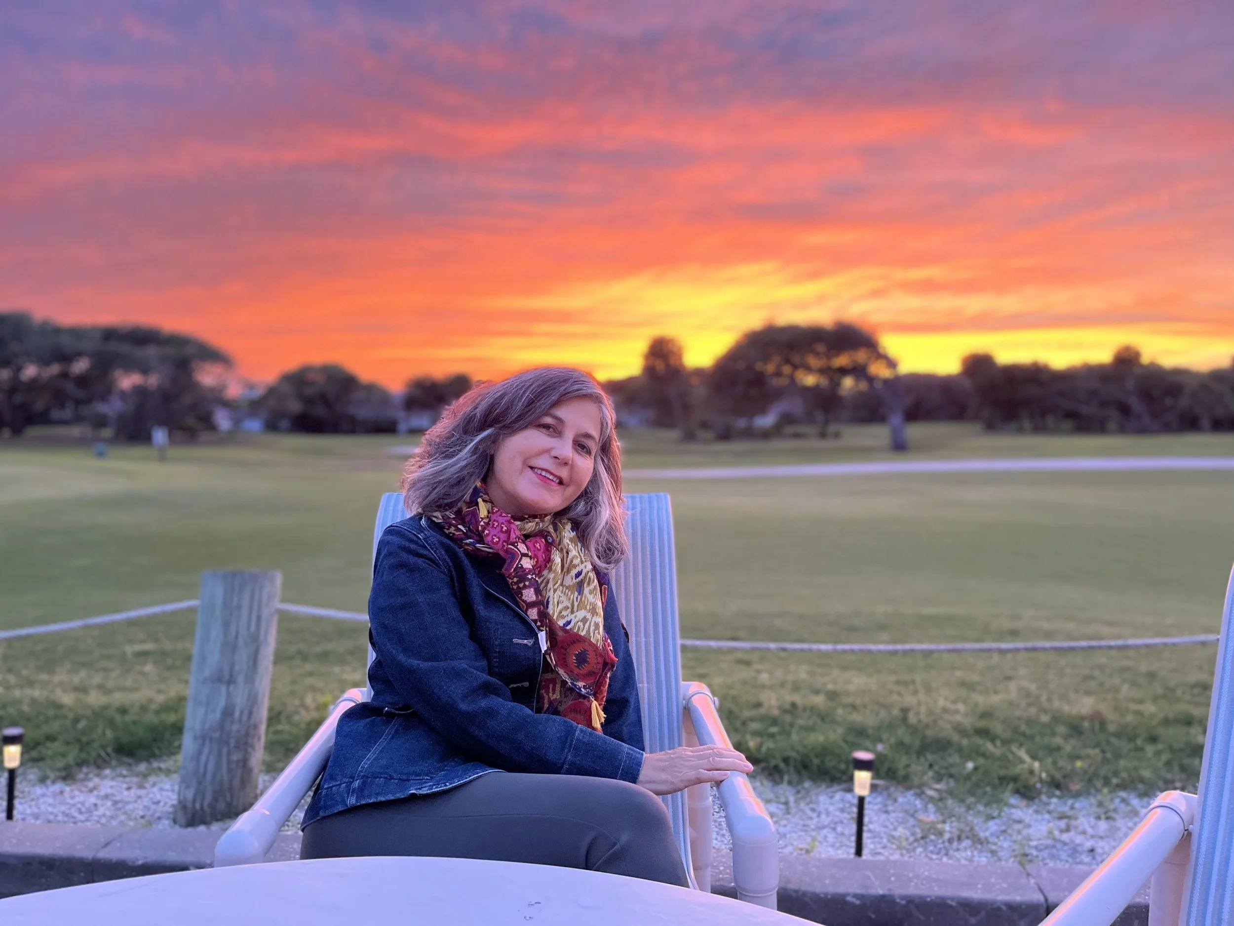 A woman with gray hair and a colorful scarf sitting on a lounge chair outdoors during sunset, with a vibrant pink, orange, and purple sky and trees in the background.