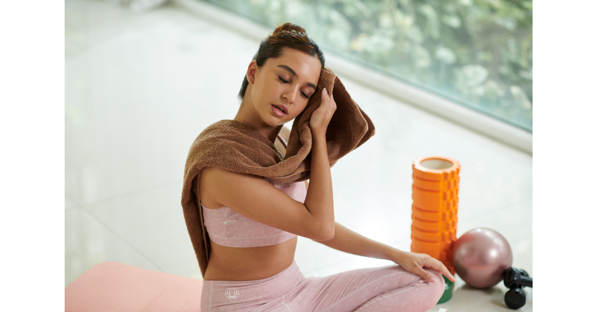 A woman in pink workout clothes sitting on a pink exercise mat, wiping sweat from her face with a towel draped over her shoulders, surrounded by foam rollers, a pink exercise ball, and a dumbbell, in a bright room near a large window.