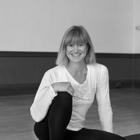 Woman with shoulder-length hair smiling, sitting on the floor in a dance studio or gym, wearing a white long-sleeve shirt and black leggings.