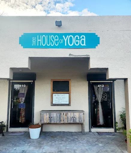 Exterior of The House of Yoga, a yoga studio, with a blue sign above the entrance. The building has two glass doors labeled 410 and 412, and a small window between them. There's a wooden bench and potted plant outside.