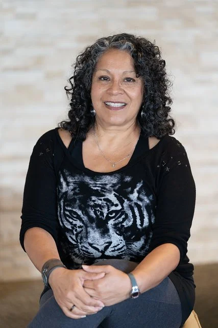 A woman with curly gray hair sitting and smiling at the camera, wearing a black shirt with a tiger face design and gray pants, in front of a neutral background.